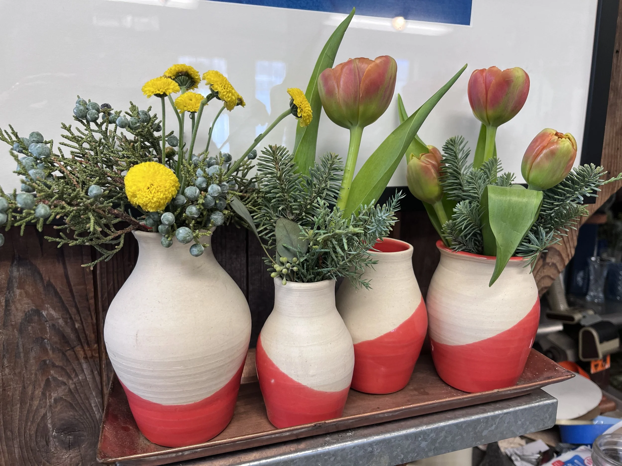 Four ceramic vases with a red and white swirl pattern, each containing different flower arrangements including yellow billy buttons and pink tulips, placed on a copper-colored tray on a wooden surface.