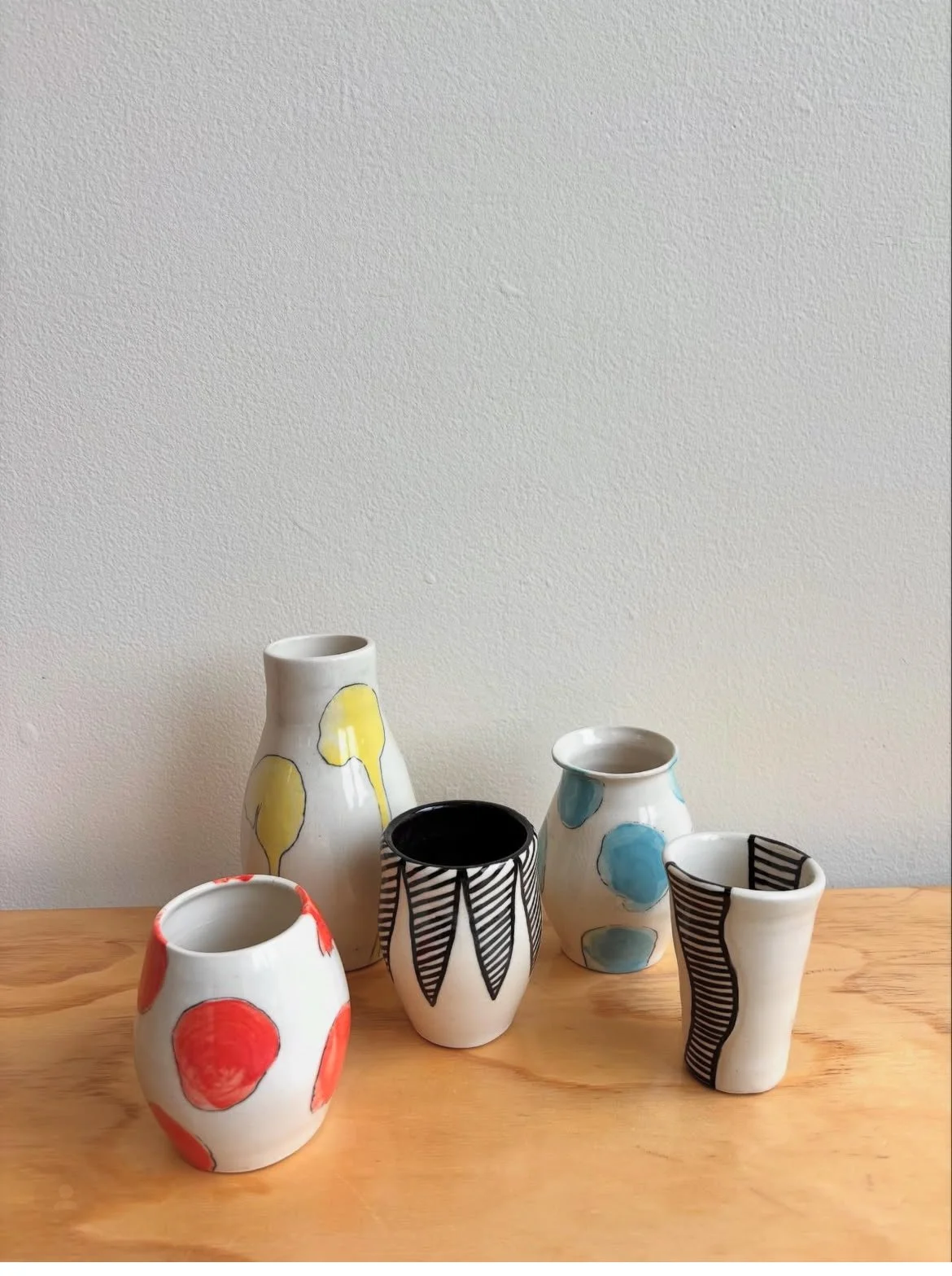 Six hand-painted ceramic vases on a wooden surface against a plain white wall, with various abstract designs and colors including red, yellow, blue, black, and white.
