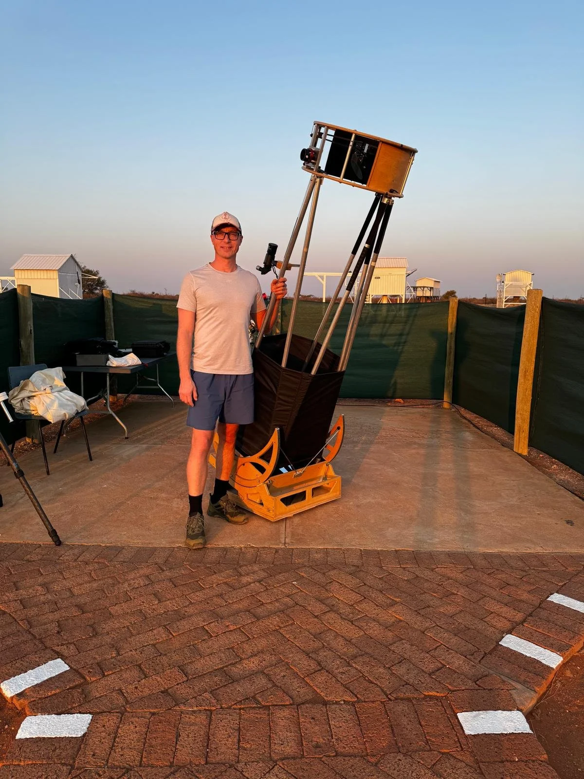 Man standing outdoors next to a large telescope mounted on a wooden base, with a fence and small buildings in the background during sunset.