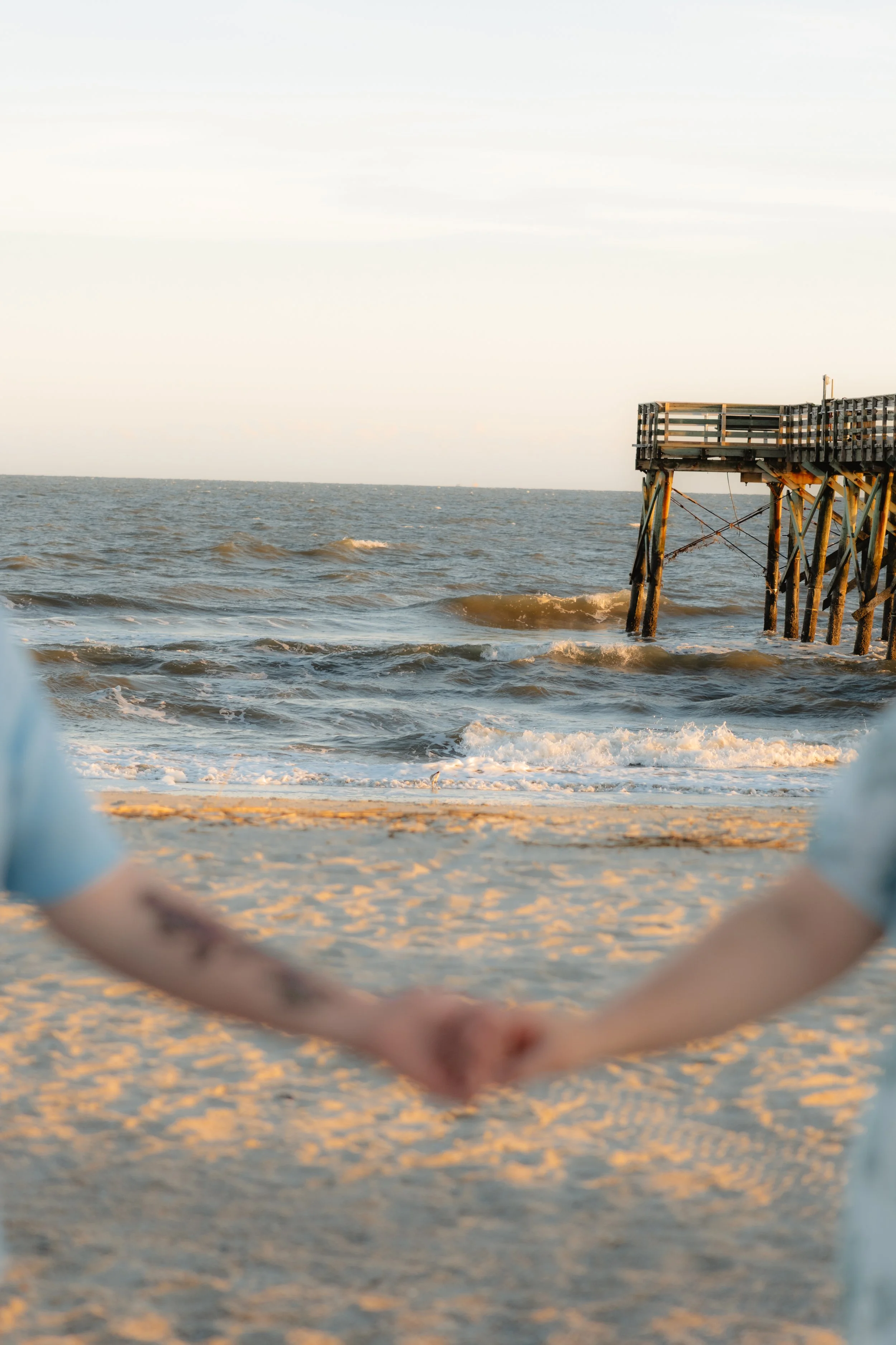 A couple holding hands on a sandy beach with a pier extending into the ocean in the background, during sunset or late afternoon.