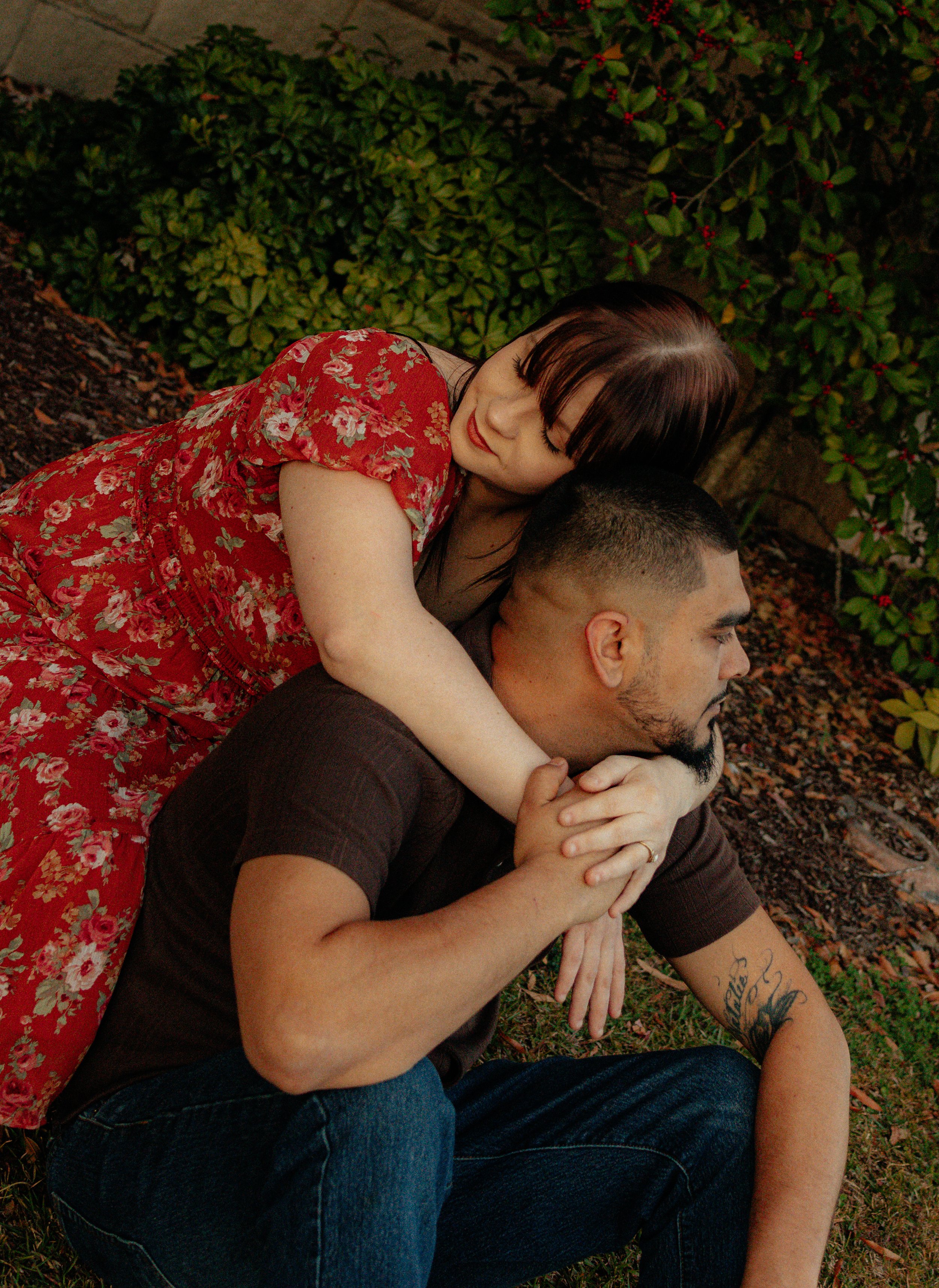 A woman in a red floral dress leaning over a man with a beard and tattoo on his arm, sitting on the ground outdoors among bushes and plants, with her arms around his shoulders.