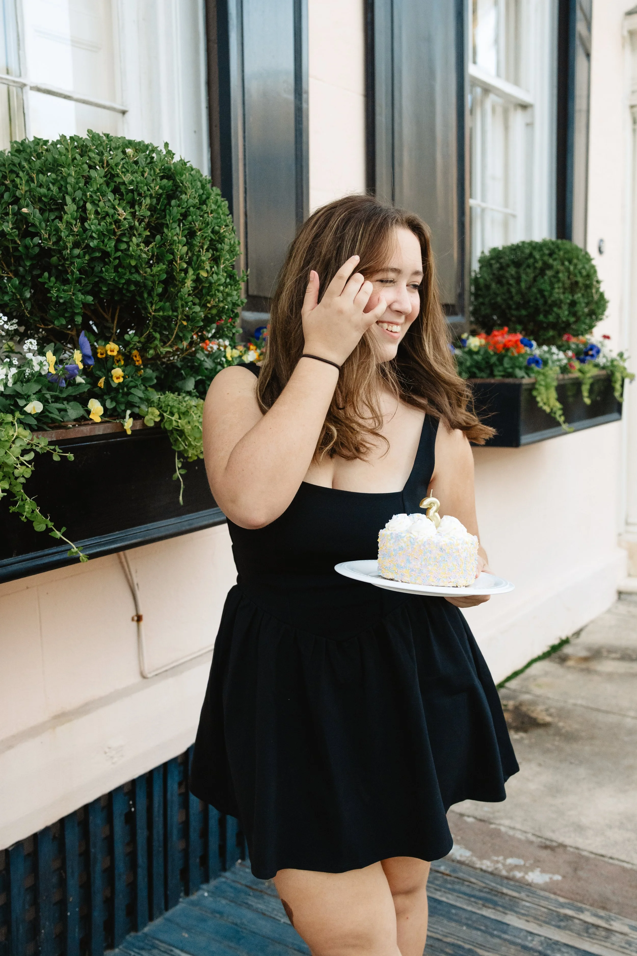 A young woman wearing a black dress holding a small birthday cake with a lit candle, smiling and standing outdoors near a building with flower boxes.