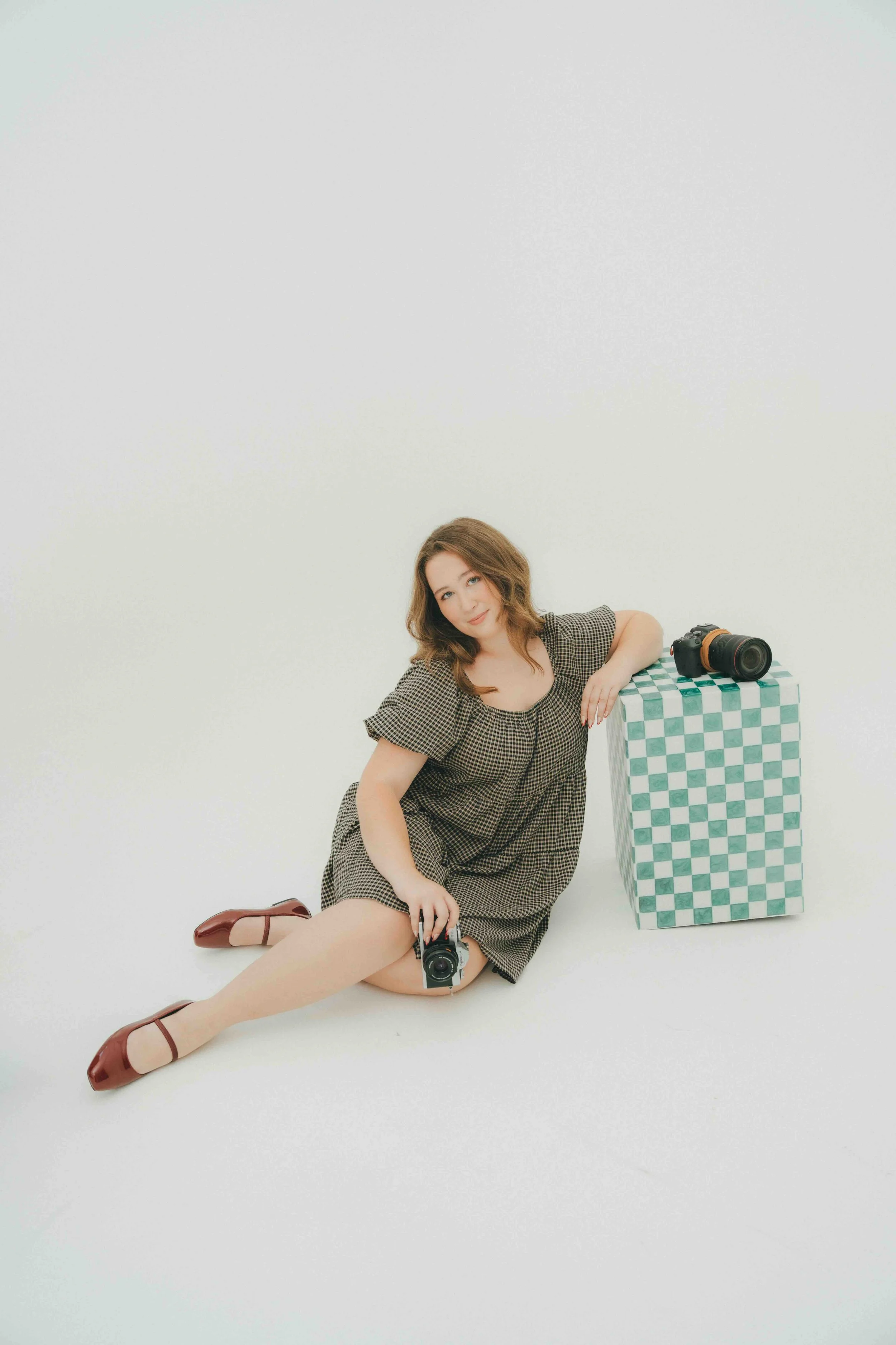 A young woman with auburn hair sitting on the floor in a photography studio, wearing a checkered dress and brown ballet flats, holding a camera, with a camera atop a checkered pedestal beside her, against a plain white background.
