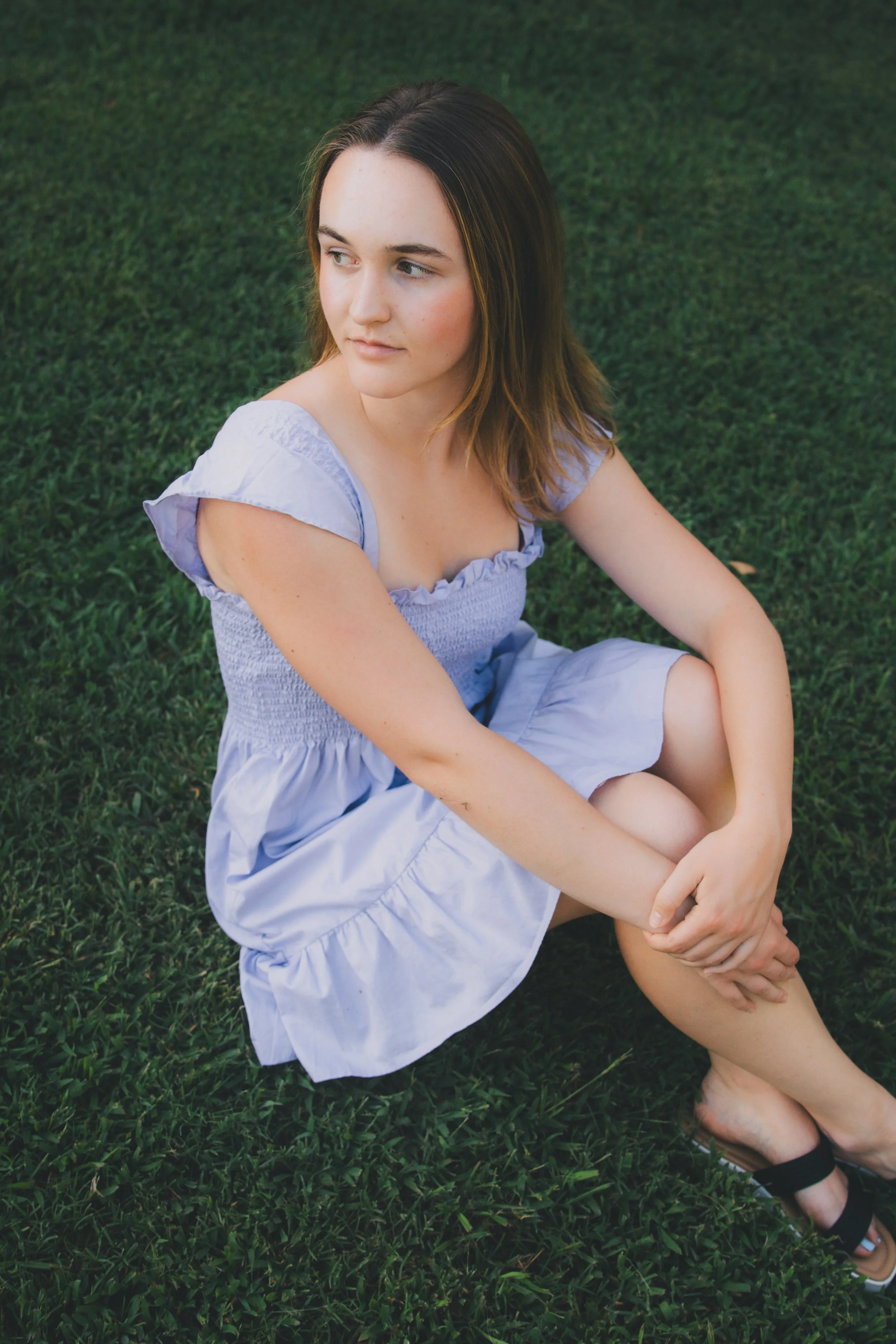 A young woman sitting on grass with her legs crossed, wearing a light purple dress, looking to her right.