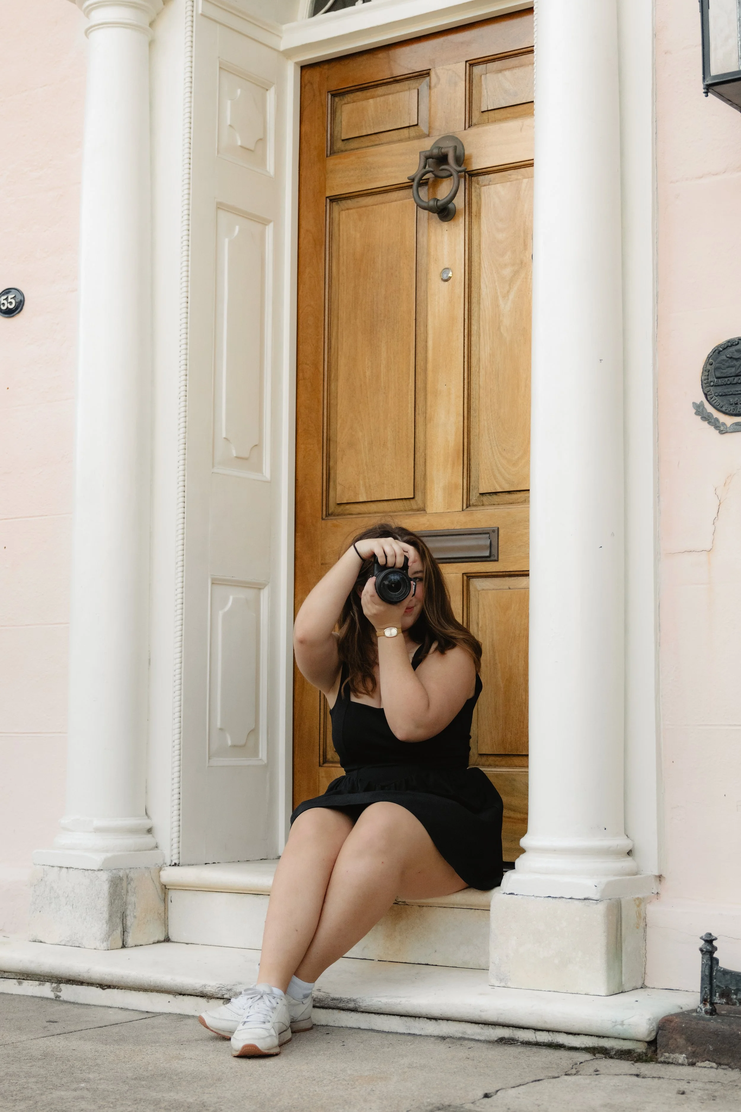 Young woman sitting on front porch steps, taking a photo of herself with a camera, wearing a black dress and white sneakers, with a wooden front door behind her.