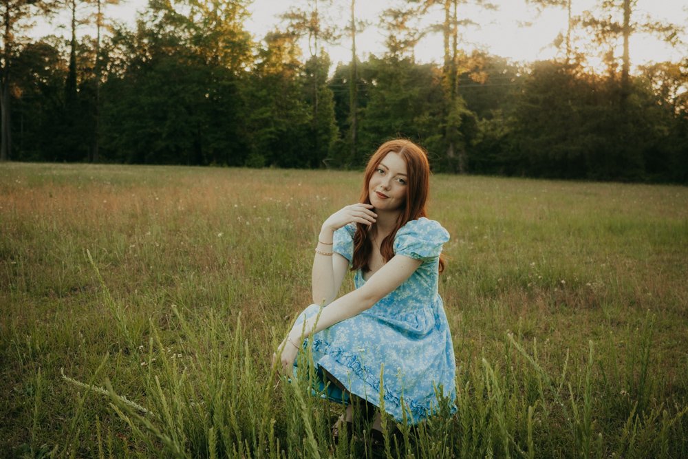 A young woman with long red hair sitting in a grassy field during sunset, wearing a blue floral dress.