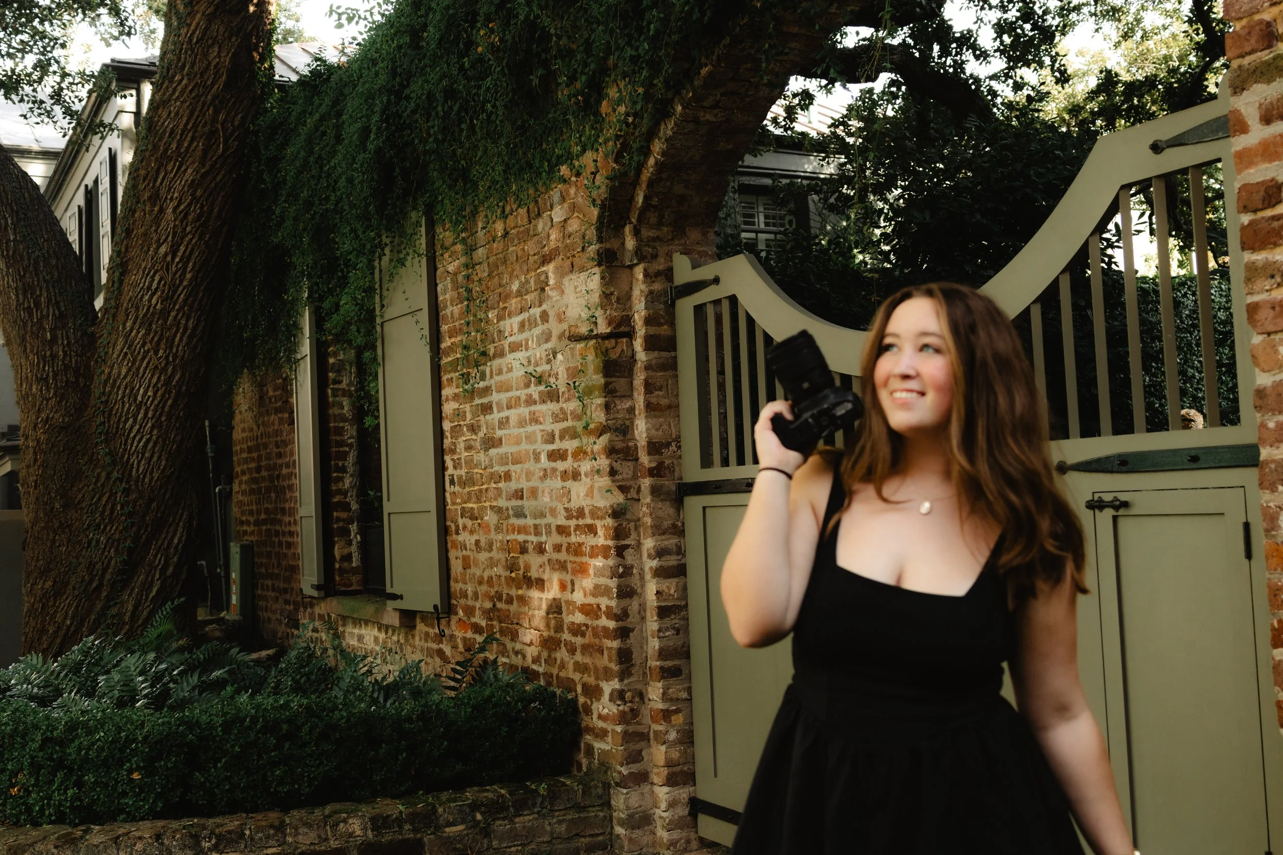 A smiling woman in a black dress holding a camera, standing outdoors near a brick wall and a large tree, with greenery and a decorative gate in the background.