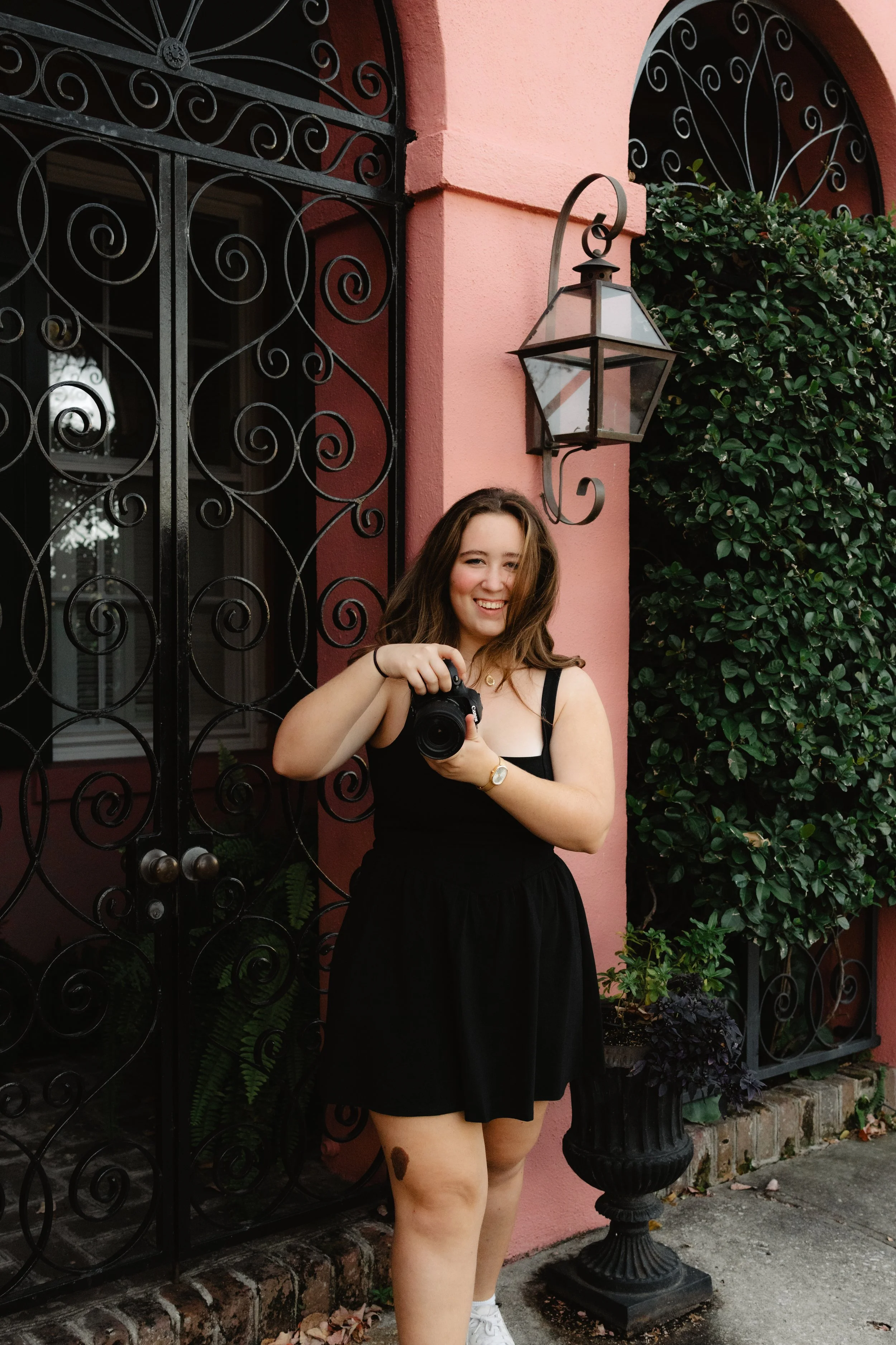 Smiling young woman in a black dress taking a photo with a camera outside a pink building with black wrought-iron gates and lush green plants.