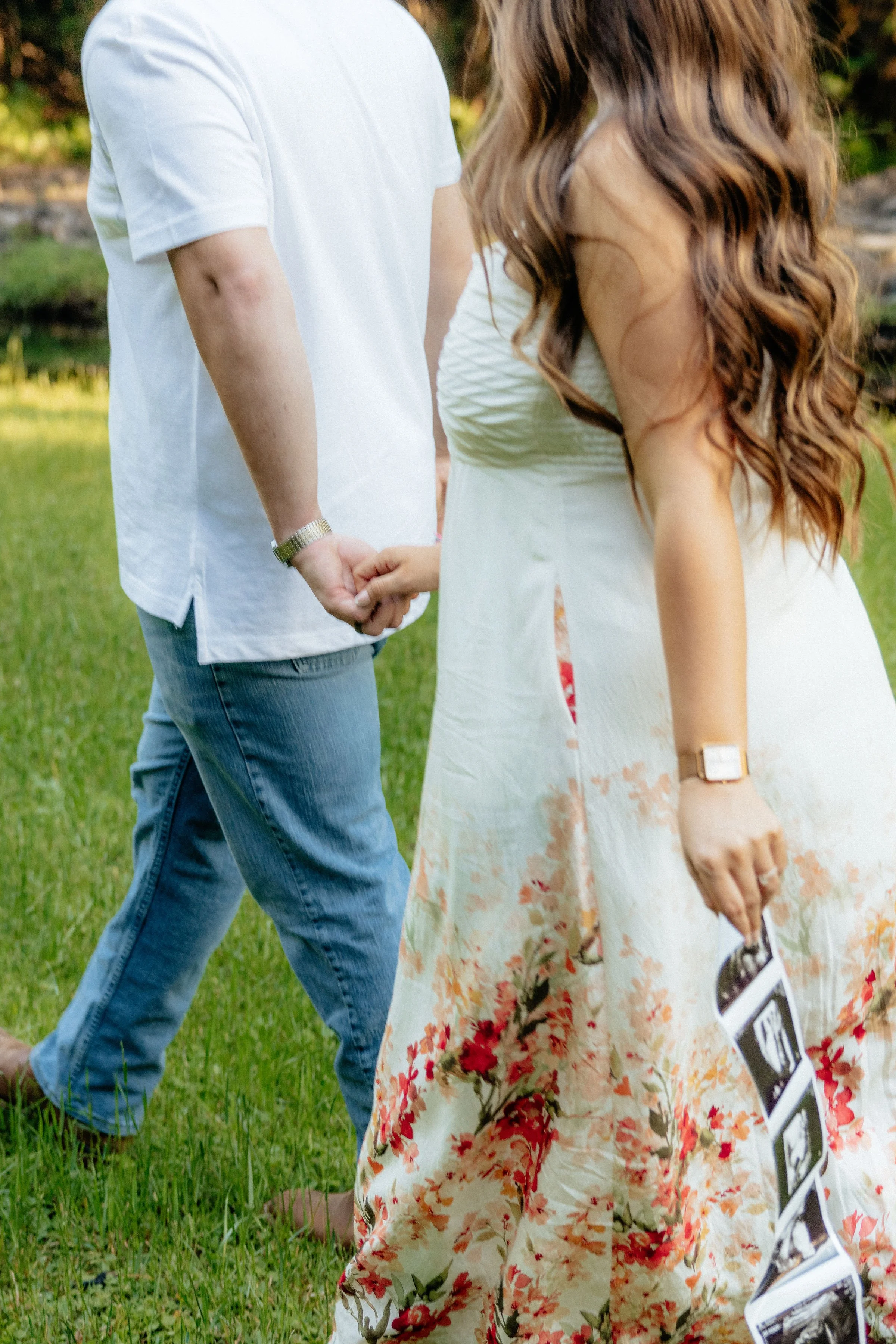 A man and woman holding hands outdoors on green grass. The woman is wearing a floral dress and the man is dressed casually in a white t-shirt and blue jeans.