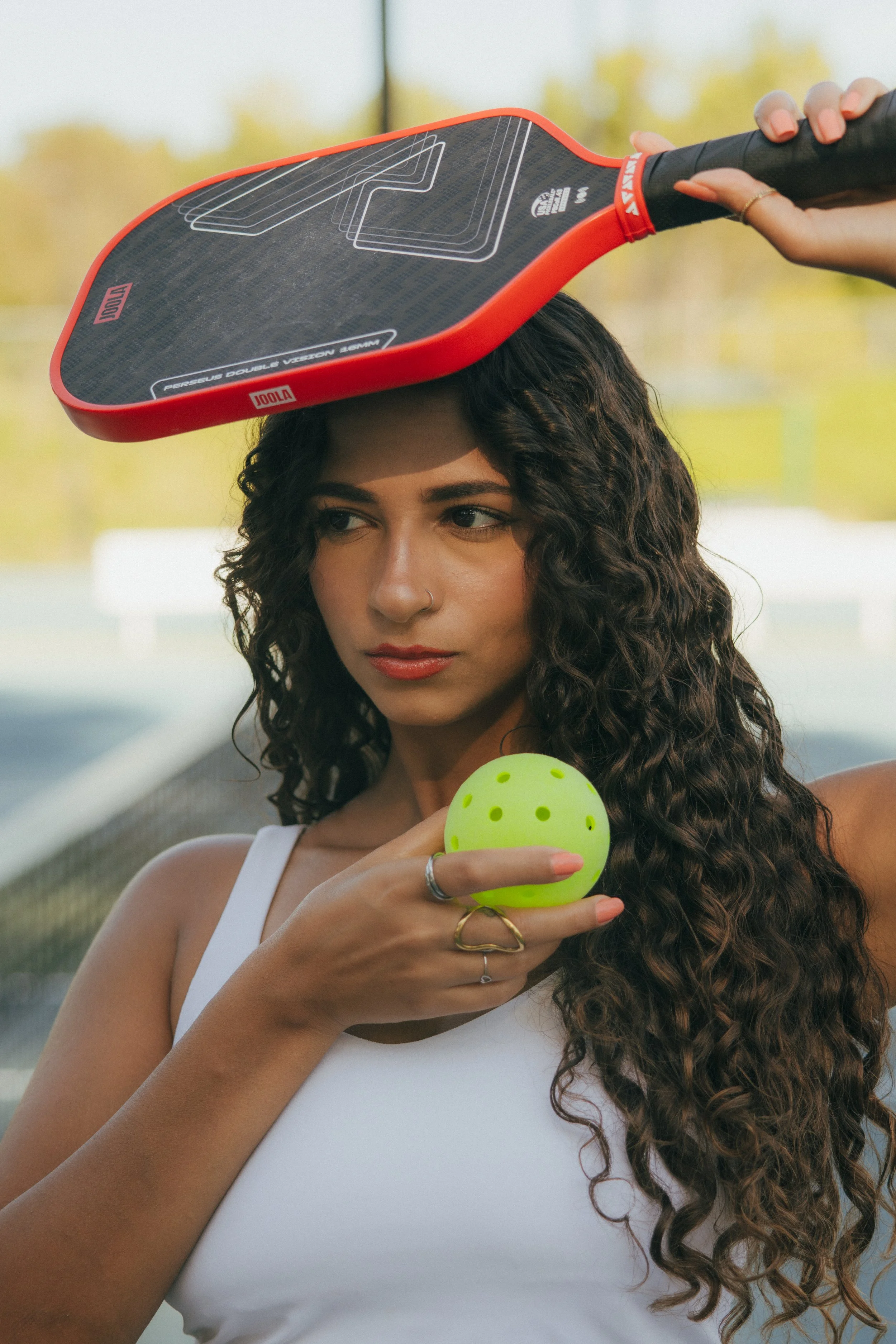 A woman with curly hair and a nose piercing holding a yellow rubber ball, standing outdoors with trees in the background. Someone is placing a paddle on her head.