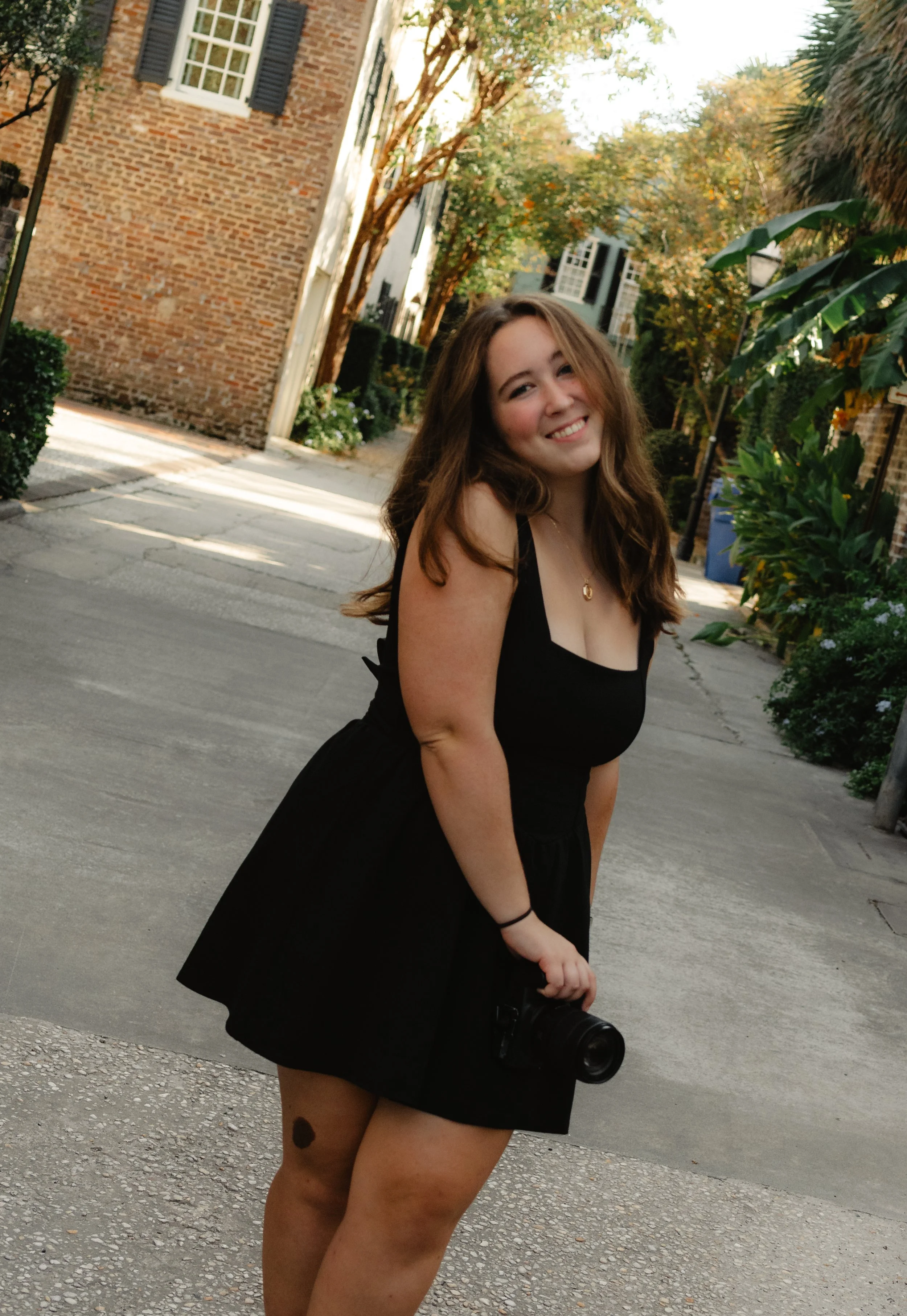 A young woman with long brown hair wearing a black dress, holding a camera, smiling, standing on a sidewalk in a suburban neighborhood with trees, shrubs, and brick houses.