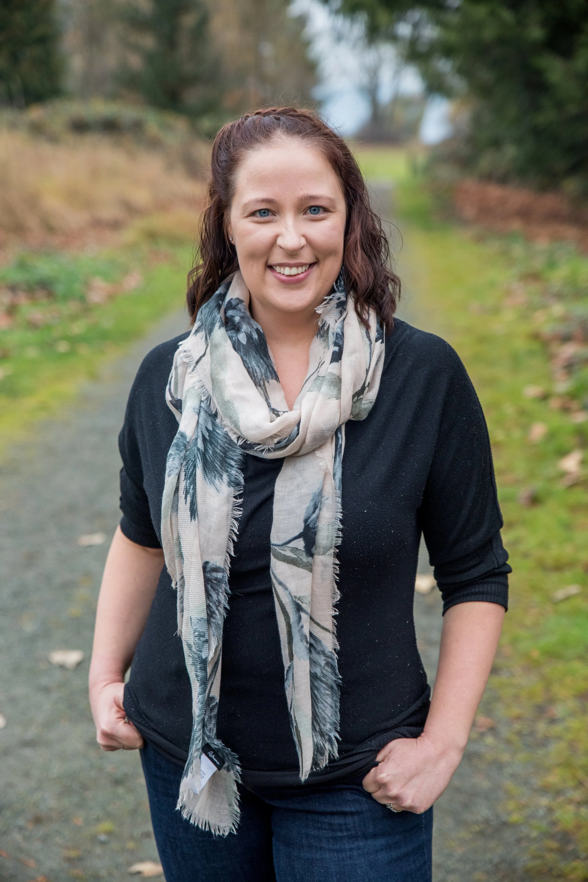 A woman with shoulder-length brown hair and blue eyes, smiling and standing outdoors on a nature trail during daytime. She is wearing a black long-sleeve top and a beige scarf with a leaf pattern. Her hands are in her pockets, and the background features green grass, trees, and a cloudy sky.