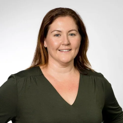 A woman with shoulder-length brown hair, wearing a dark green V-neck top, smiling against a plain white background.