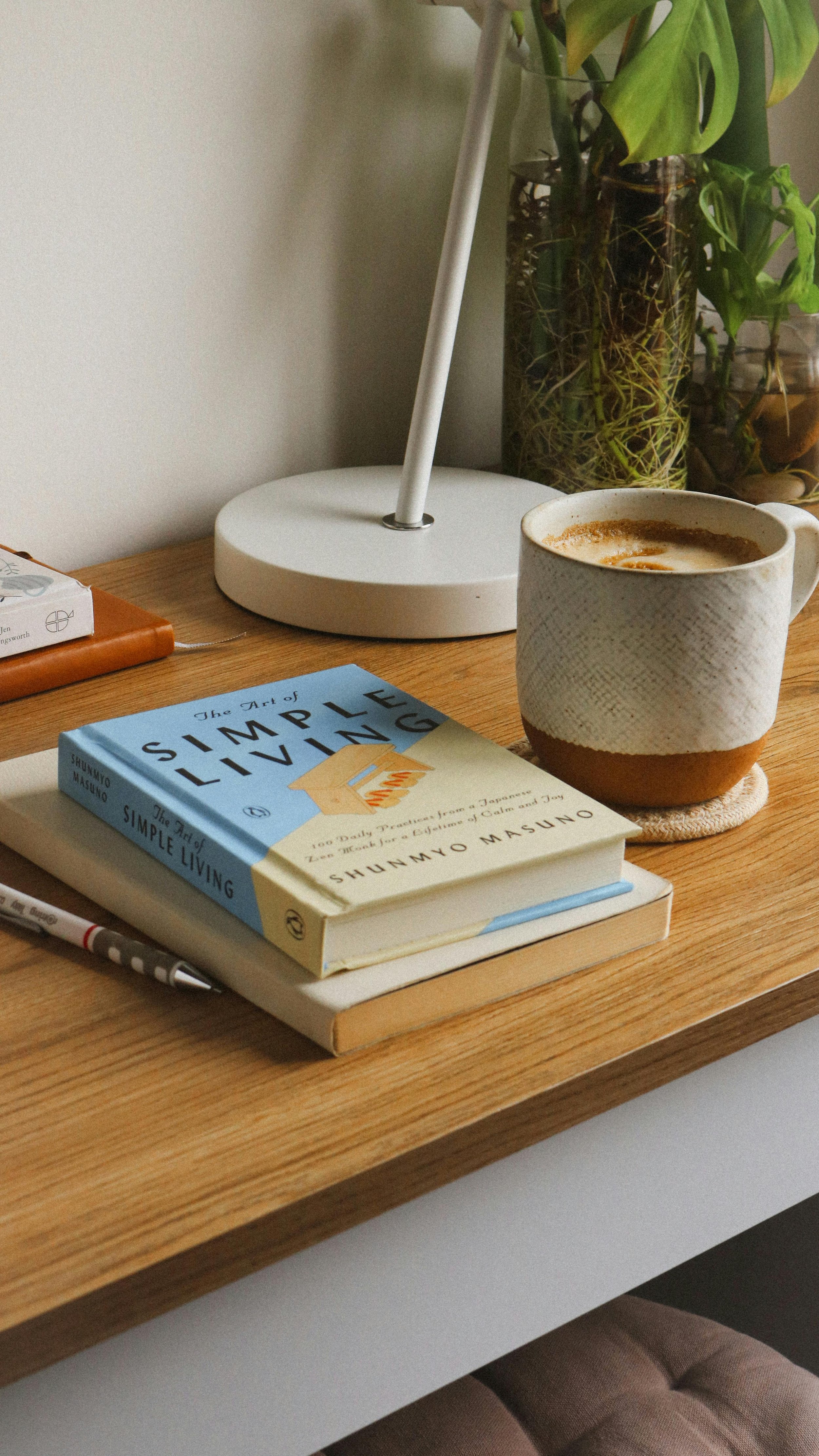 A wooden desk with a book titled 'The Art of Simple Living' by Shunmyo Masuno, a notebook, a pen, a white lamp, and a ceramic mug with coffee, with potted plants in the background.