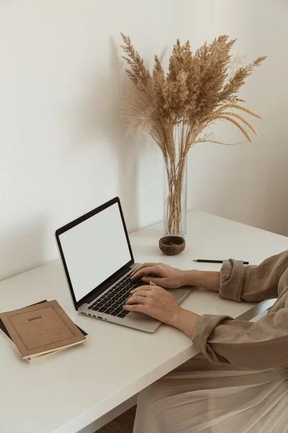 Person typing on a laptop at a white desk with a notebook, pen, and a vase of dried pampas grass in the background.