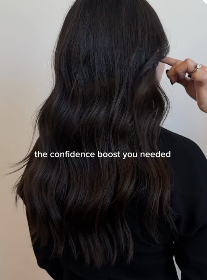 A woman with long, wavy, dark brown hair, seen from behind, touching her hair with her right hand, wearing a black top, with the caption 'the confidence boost you needed' over the image.