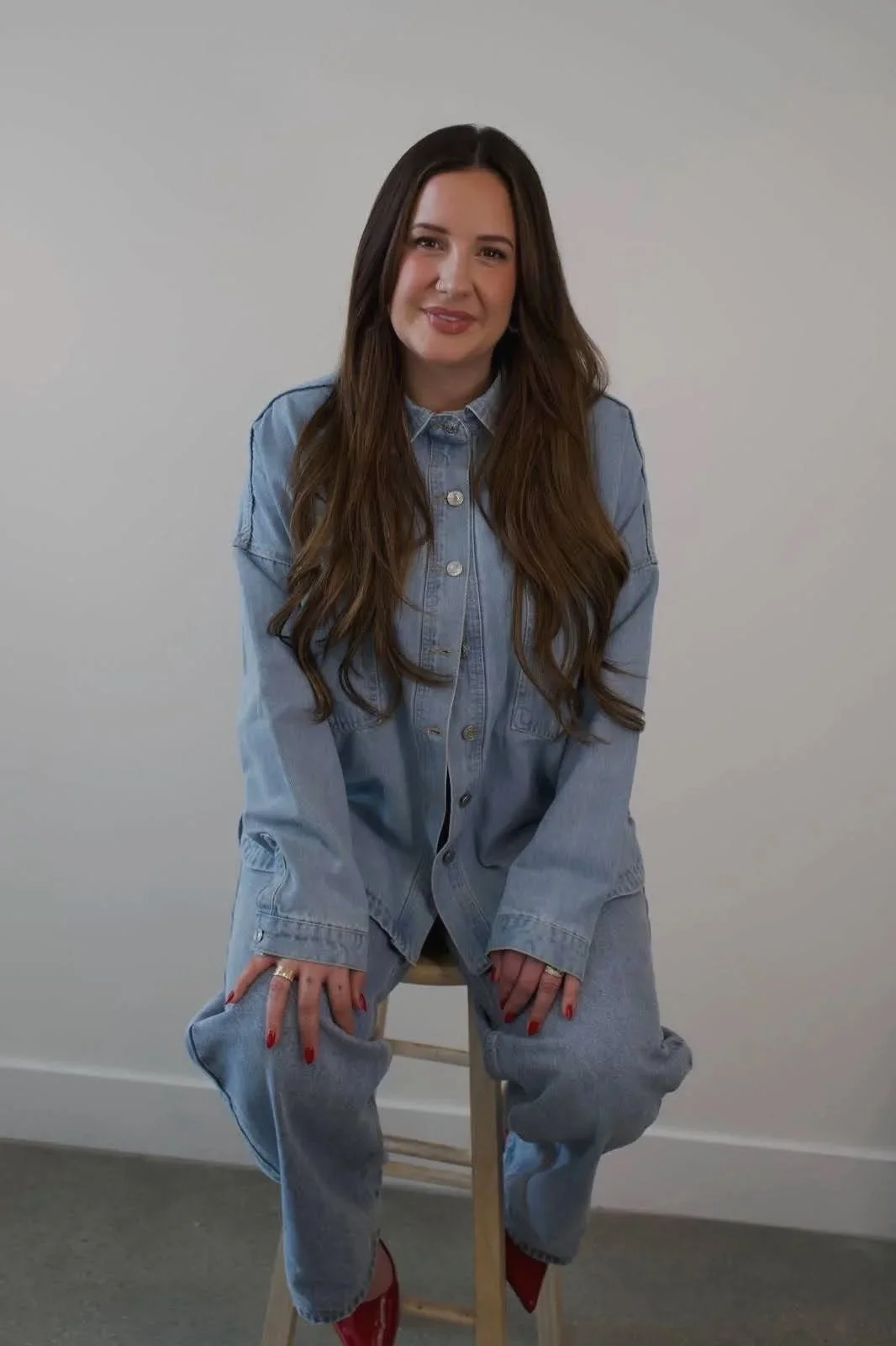 A woman with long brown hair wearing a light blue denim shirt and matching jeans sitting on a wooden stool against a plain white background.