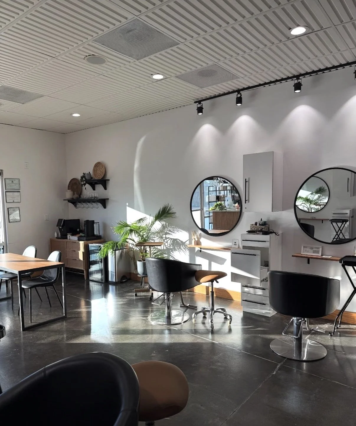 Interior view of a modern salon or hair studio with styling chairs, mirrors, and a coffee station, featuring sunlit space, plants, and minimalist decor.