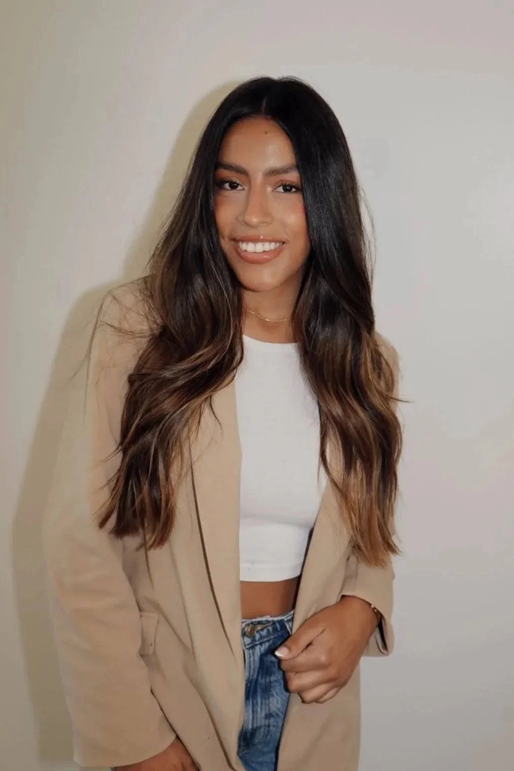 A young woman with long wavy dark brown hair and medium skin tone, smiling, wearing a beige blazer over a white crop top, blue jeans, and a small gold necklace, standing against a plain light-colored wall.