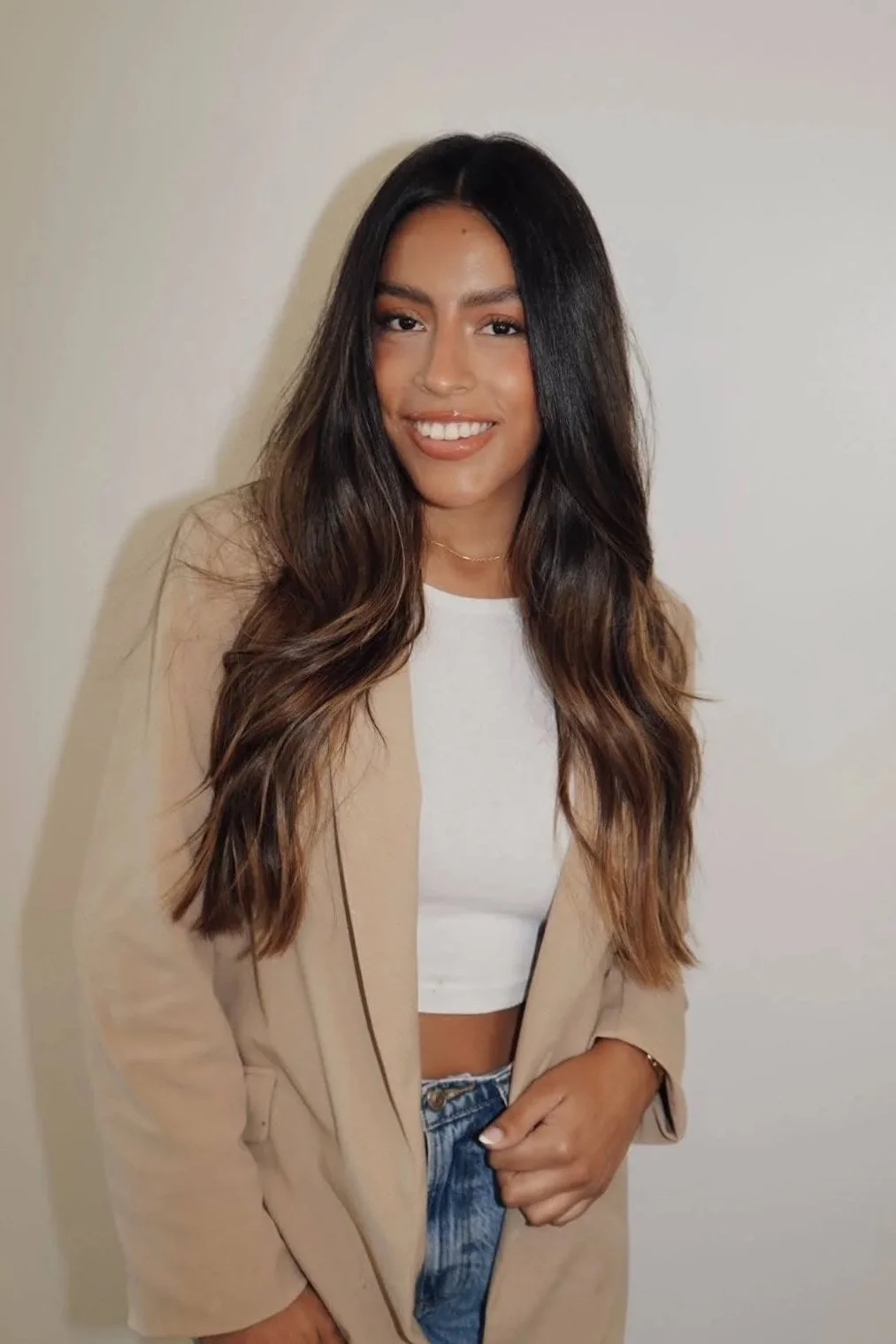 A young woman with long, wavy dark hair and a warm smile posing against a plain white wall. She is wearing a beige blazer, a white crop top, and blue jeans, with subtle makeup and a delicate necklace.