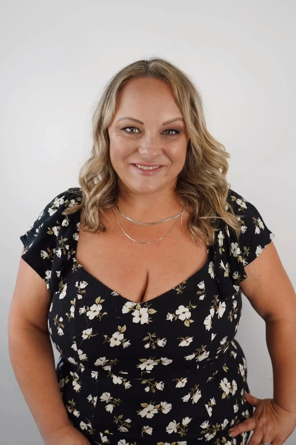 A woman with shoulder-length wavy blonde hair wearing a black floral dress and layered necklaces standing in front of a plain white wall.