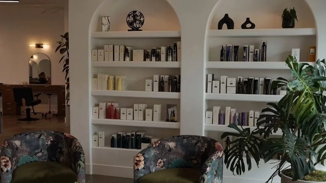 Interior of a modern room with white arched bookshelves filled with books and decorative objects, two floral upholstered chairs, and a large leafy potted plant.