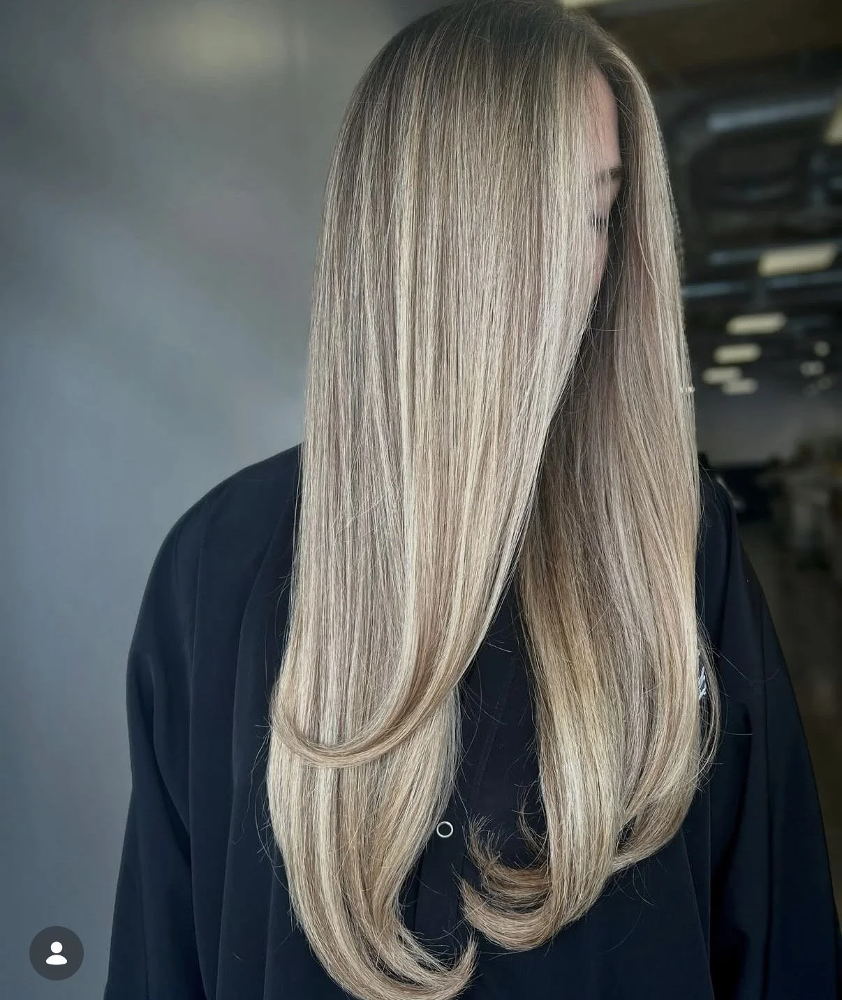 A woman with long, blonde, straight hair covering her face, wearing a black jacket in an indoor setting.