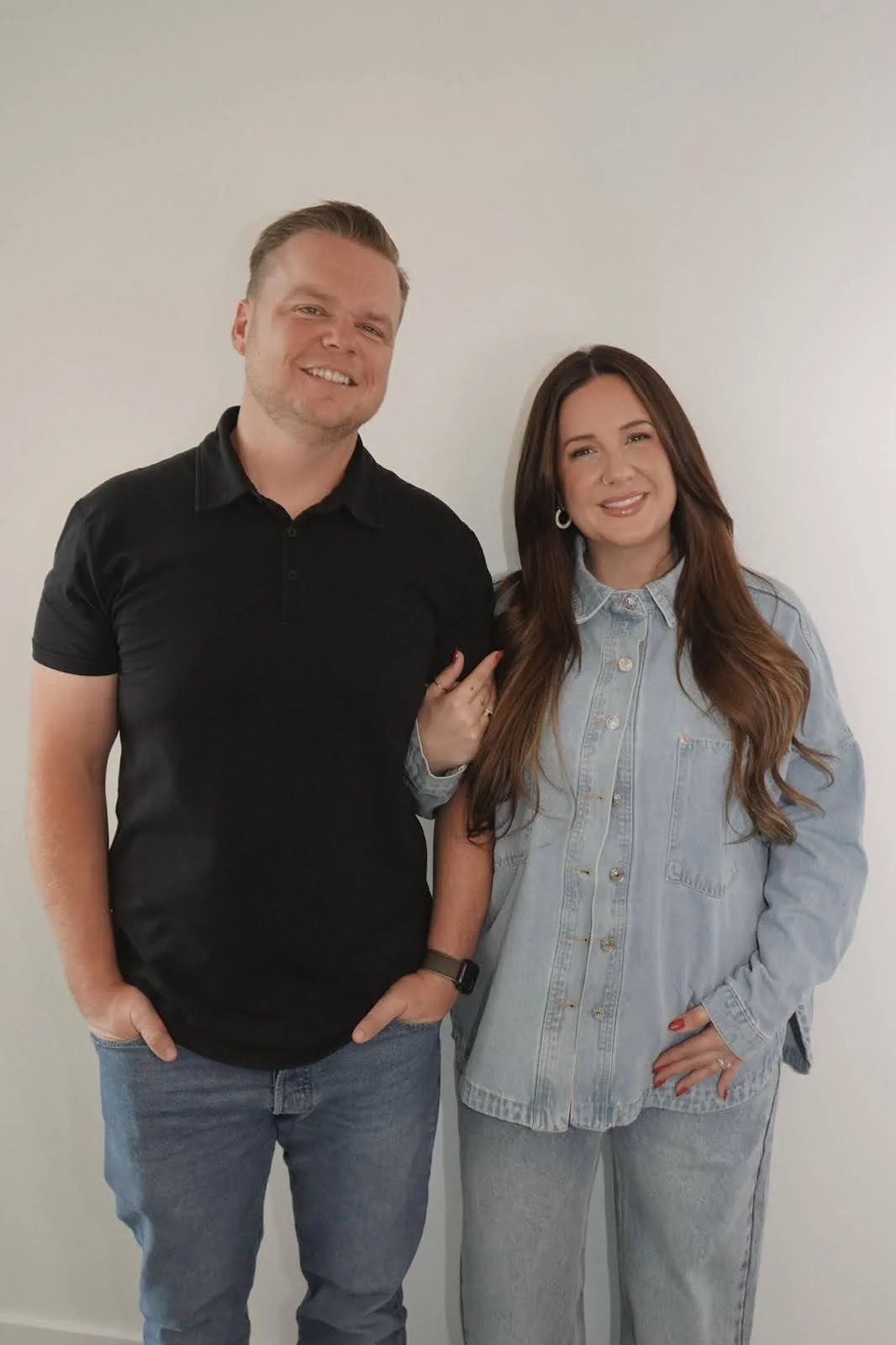 A man and woman (the owners) standing together in front of a plain white wall, smiling at the camera. The man is wearing a black polo shirt and jeans, the woman is wearing a light denim jacket and jeans.