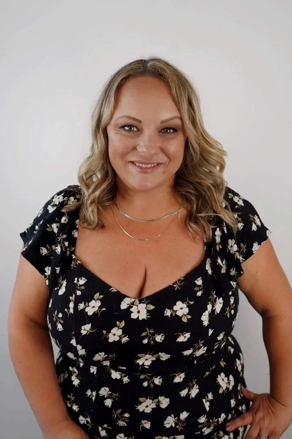 A woman with shoulder-length curly blonde hair, wearing a black floral dress and silver jewelry, smiling at the camera against a plain white background.