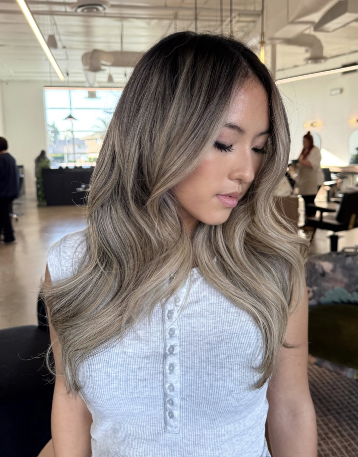 A young woman with long, wavy, blonde hair is looking down. She is wearing a light grey, buttoned sleeveless top.