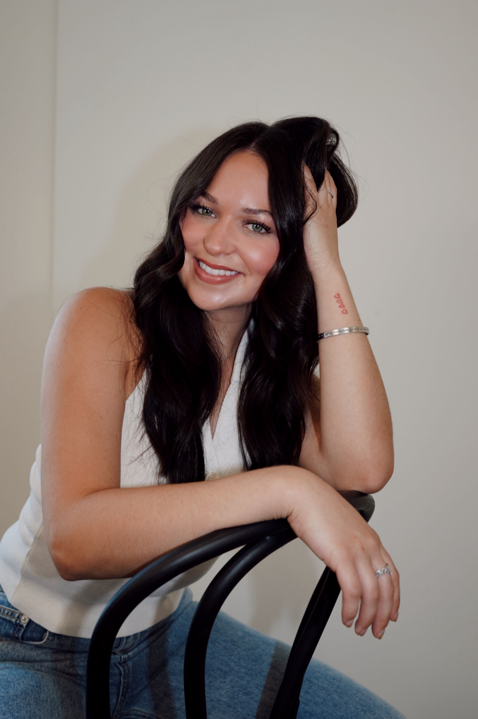 A woman with long dark wavy hair, smiling, wearing a white sleeveless top and blue jeans, sitting on a black chair, resting her arm on it, with a plain beige wall background.