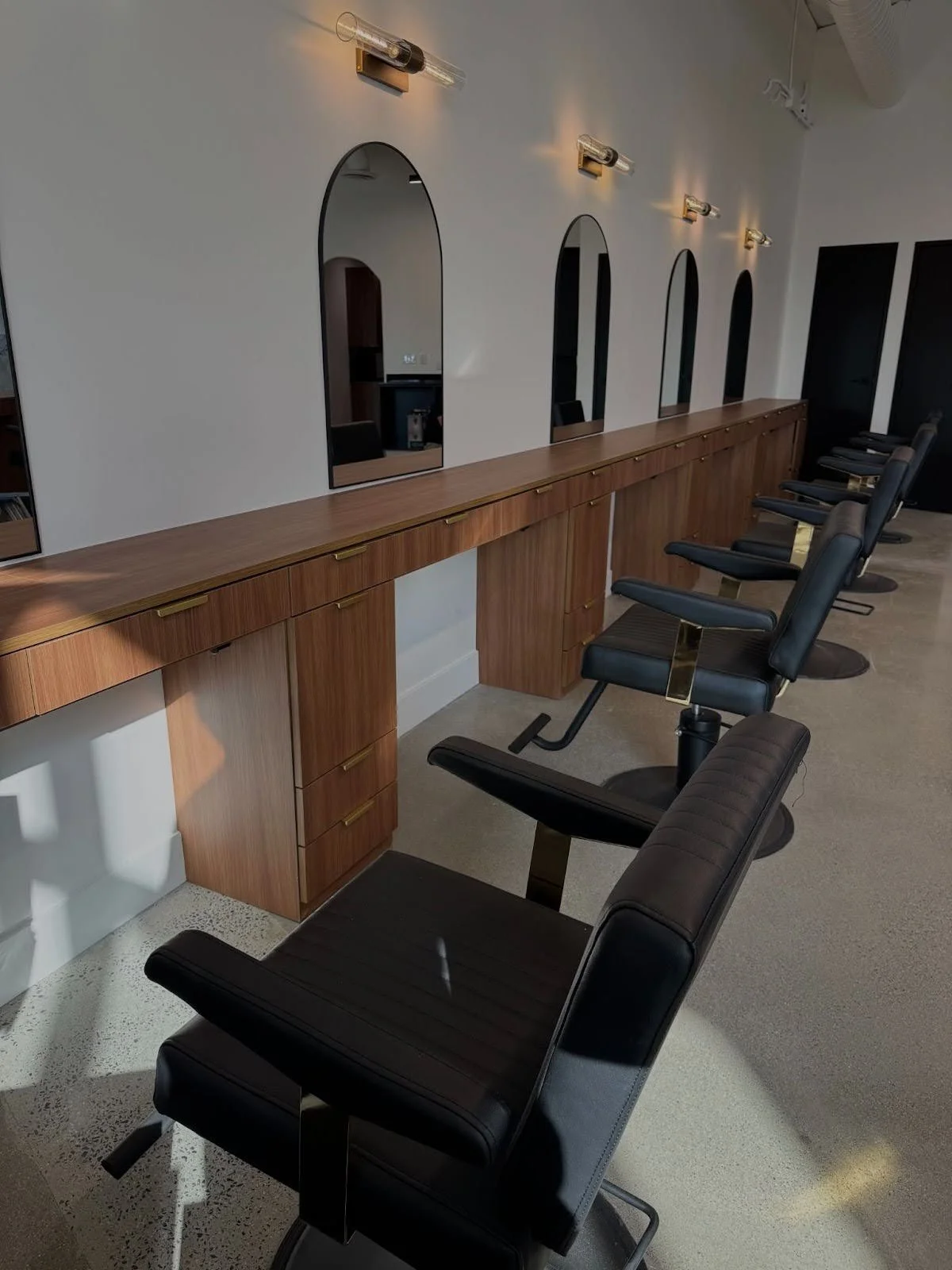 A row of black salon chairs positioned in front of a long wooden counter with five mirrors in a modern salon interior.