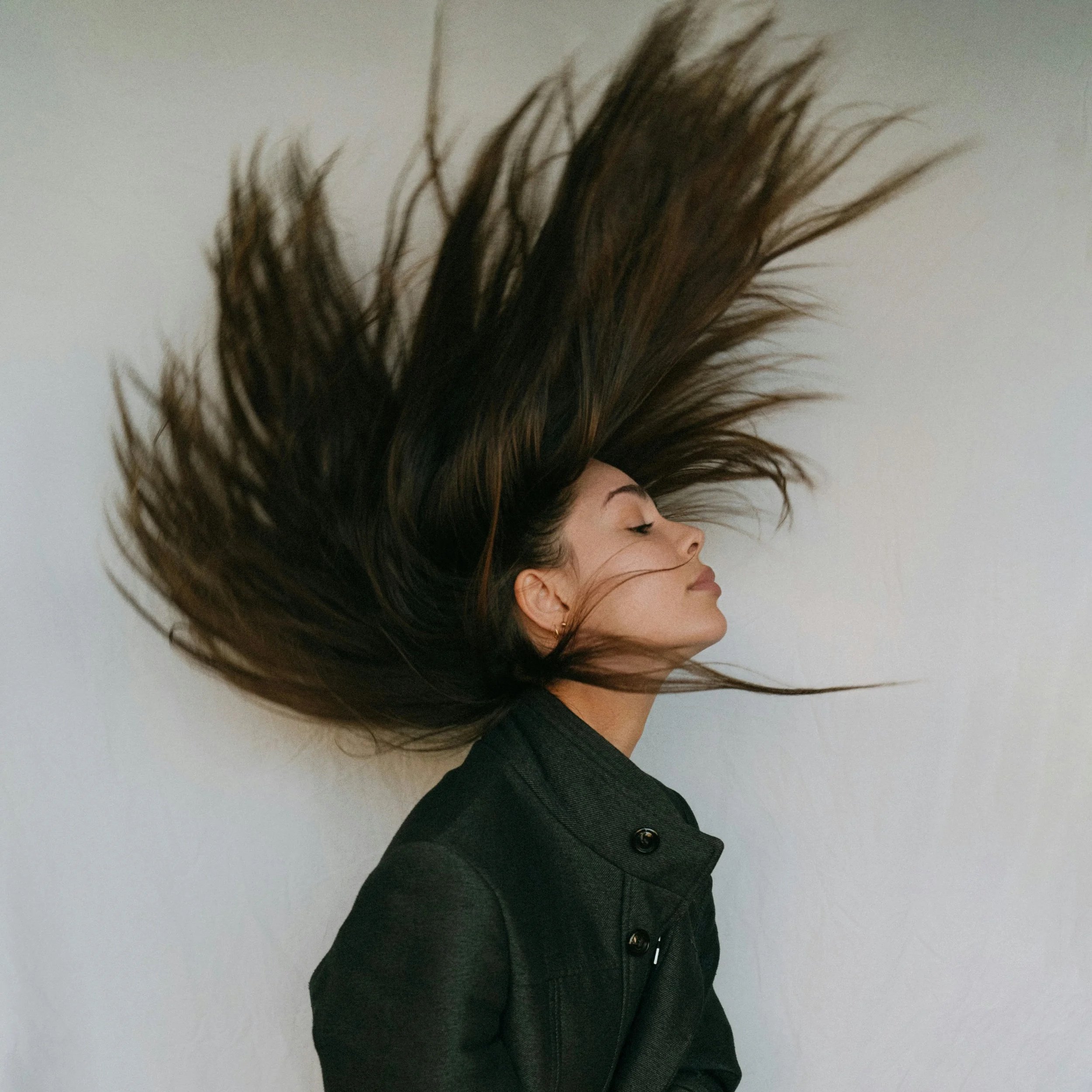 A woman with long dark hair extensions flying upward against a plain background, wearing a black jacket with a high collar.