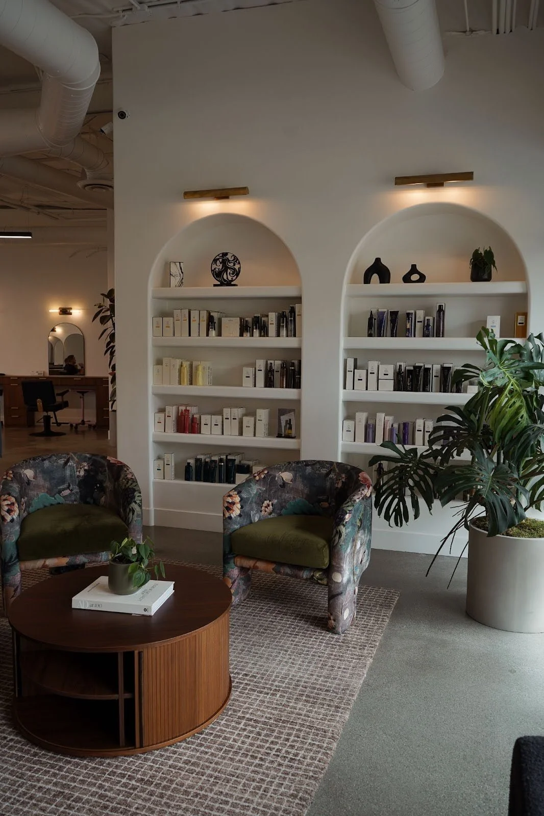 A modern lounge area with two floral patterned armchairs with green cushions, a wooden coffee table with a potted plant and books, surrounded by a beige carpet and a large potted plant nearby. Behind, white shelves built into arched wall niches hold decorative vases and books, illuminated by wall-mounted lights.