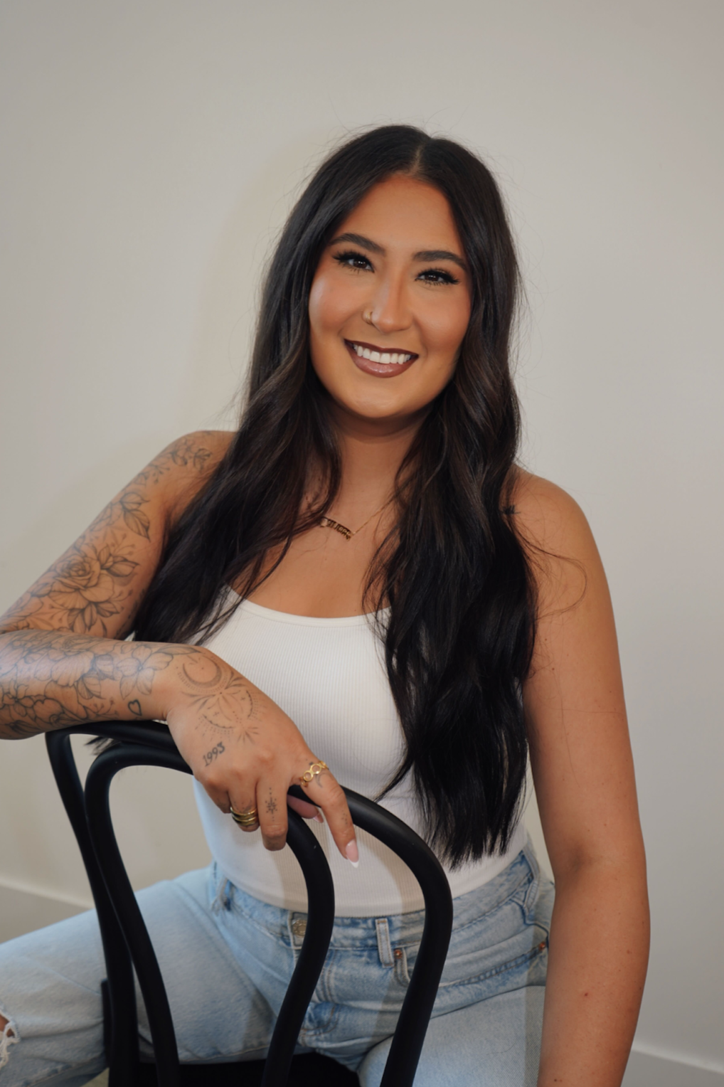 A smiling woman with long dark hair, tattoos on her arm, wearing a white tank top and light jeans, sitting on a black chair against a plain beige wall.