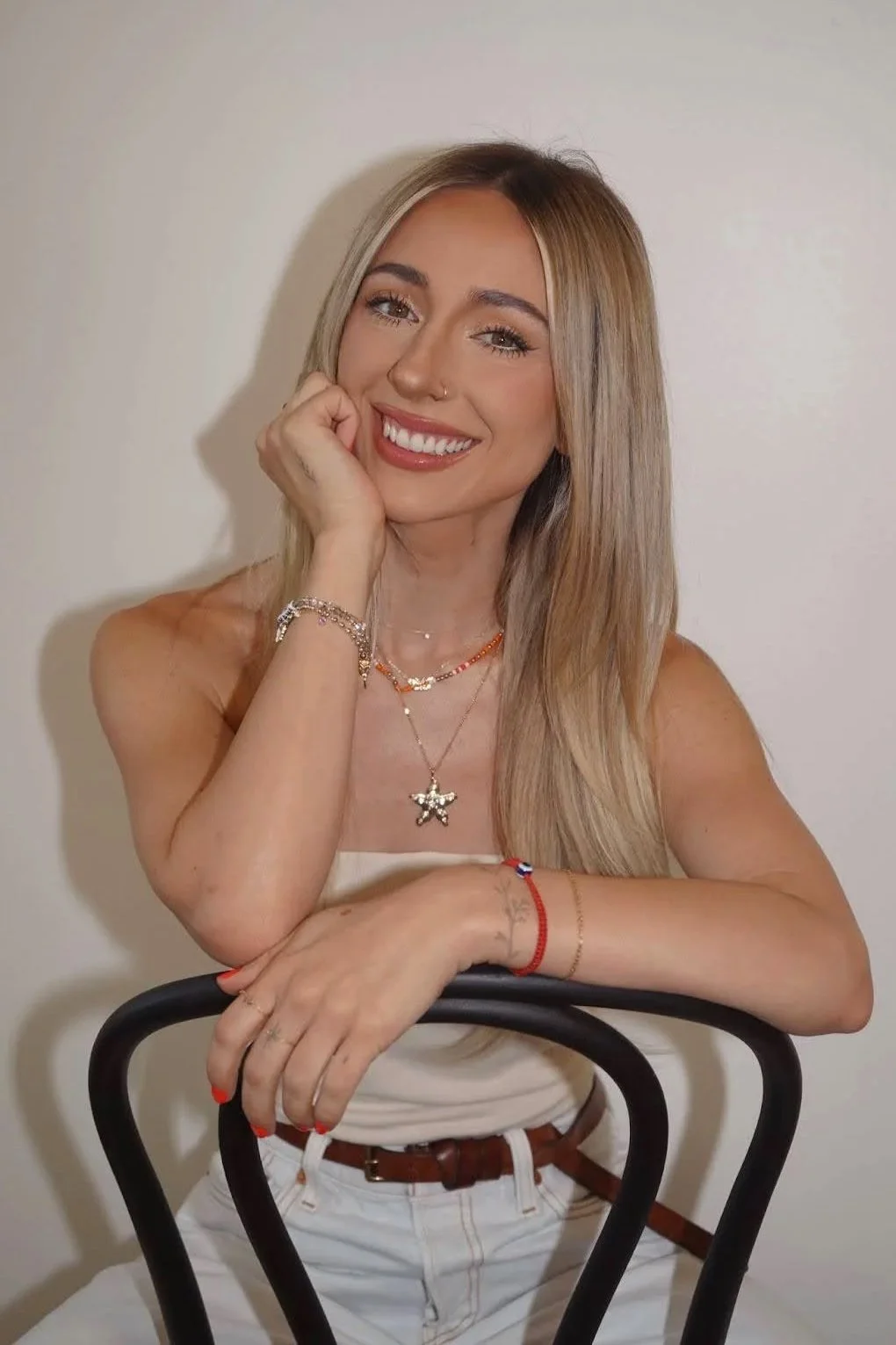 A smiling woman with long blond hair, wearing a white top and jewelry, sitting with her arm resting on a black chair, posing against a plain white wall.