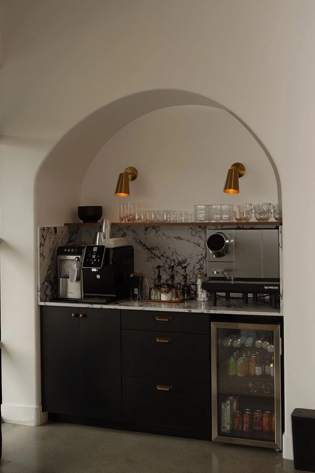 A small coffee station with black cabinets, a marble countertop and backsplash, a coffee machine, glassware, and a mini fridge with beverages inside.