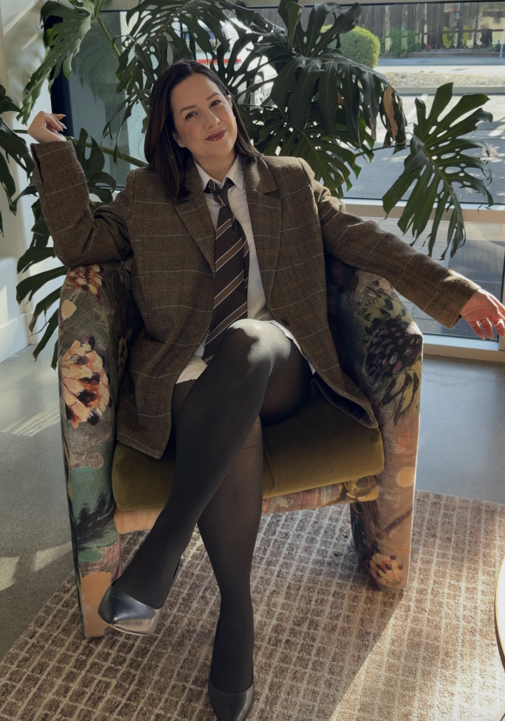 Owner and Stylist Jessica sitting in a floral armchair near a large window, wearing a brown blazer, a white shirt, a striped tie, black stockings, and metallic silver heels, with a large plant behind her.