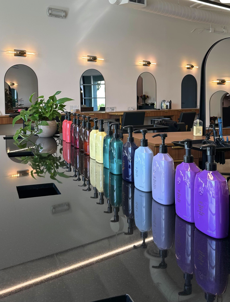 Colorful Orbie bottles of hair care products arranged on a black countertop in a salon with a large mirror, a plant, and salon chairs in the background.