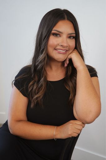 A woman with long dark hair and a nose piercing, smiling and resting her chin on her hand, sitting against a plain light gray background.