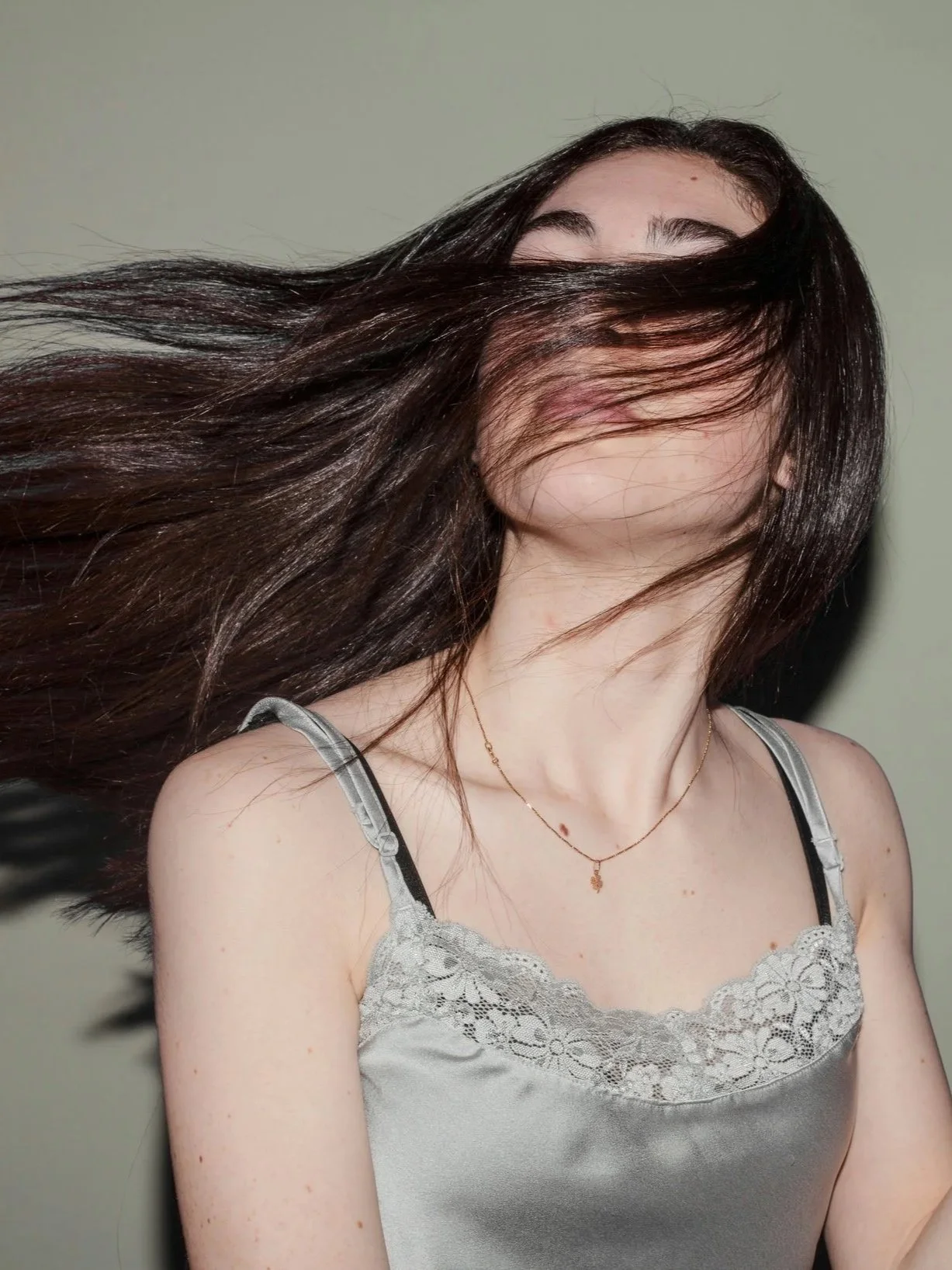 A woman with long dark brown hair, wearing a silver satin camisole with lace trim and a delicate gold necklace, her hair flowing across her face.