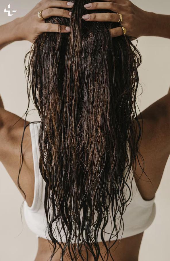 A person with long, wet, wavy dark hair holding their head with both hands, wearing a white tank top and gold rings.