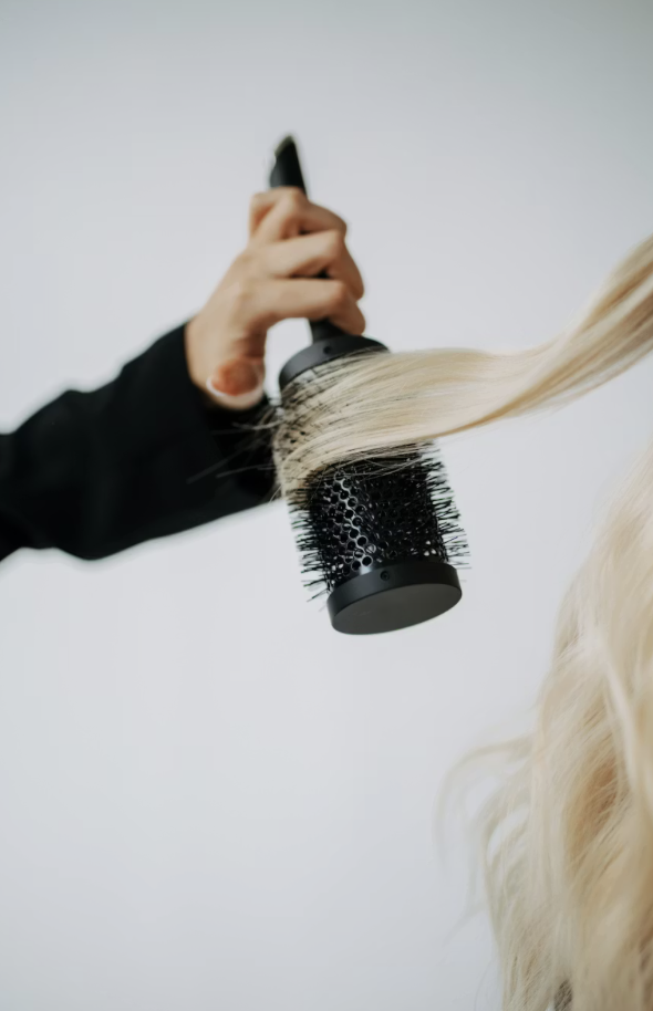 Person blow-drying long, blonde hair with a round brush.