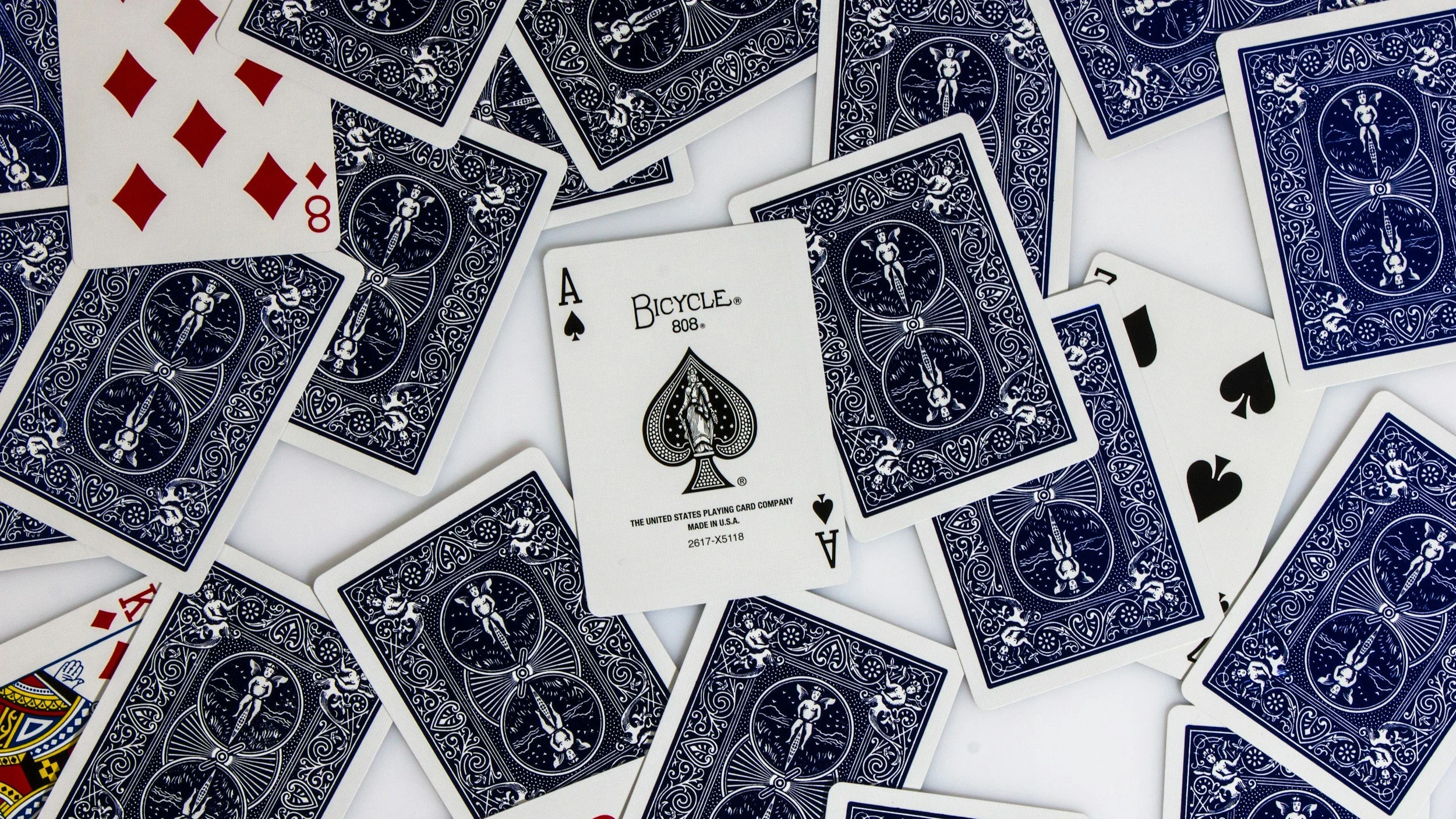 Scattered blue and white playing cards on a white background, including an Ace of Spades and an Ace of Diamonds, with a prominent Bicycle brand Ace of Spades card in the center.