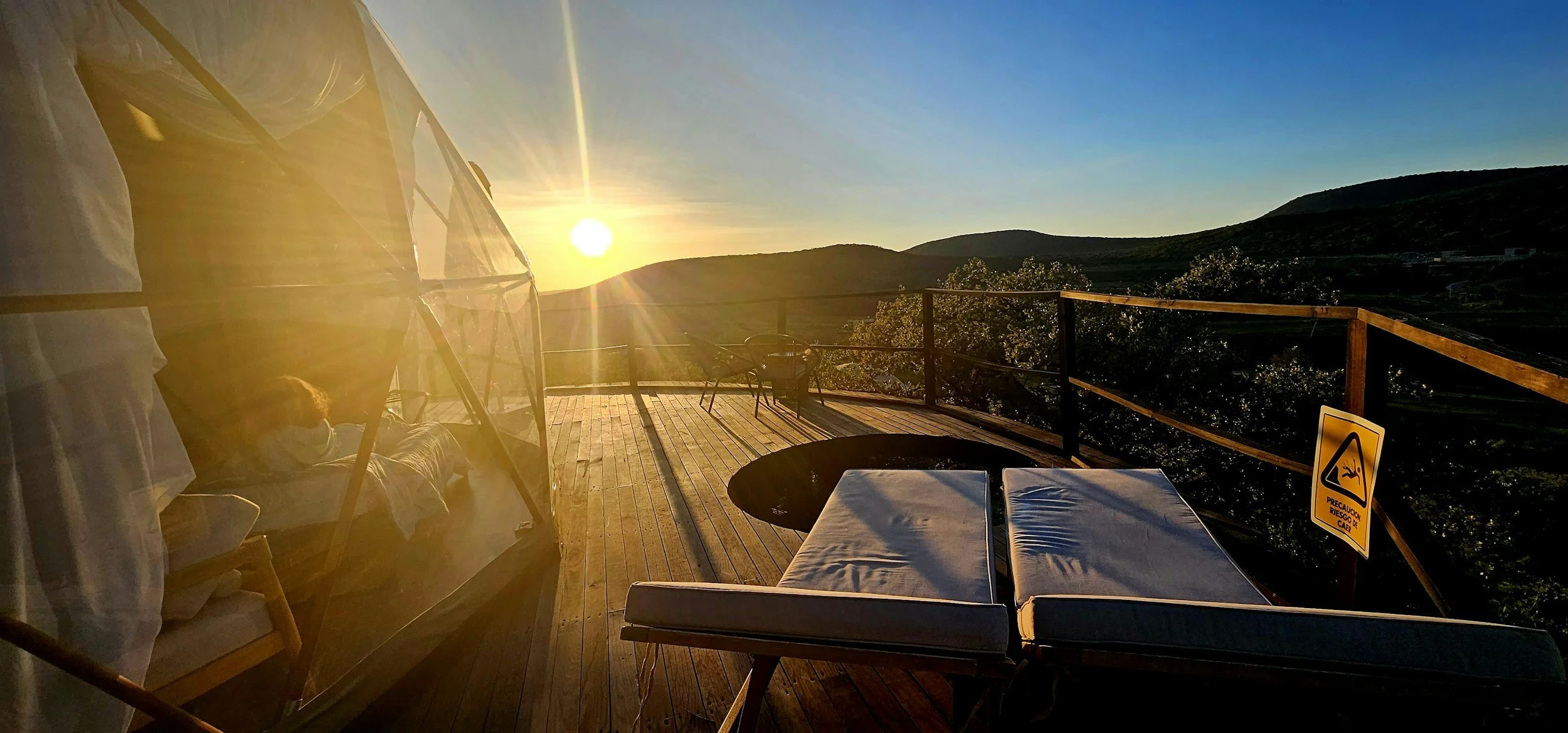 A scenic outdoor wooden deck overlooking a hilly landscape at sunset. The deck has lounge chairs, a table, and a person relaxing on a bed under a canopy. There's a warning sign on the railing, and the sun is low on the horizon casting warm light.