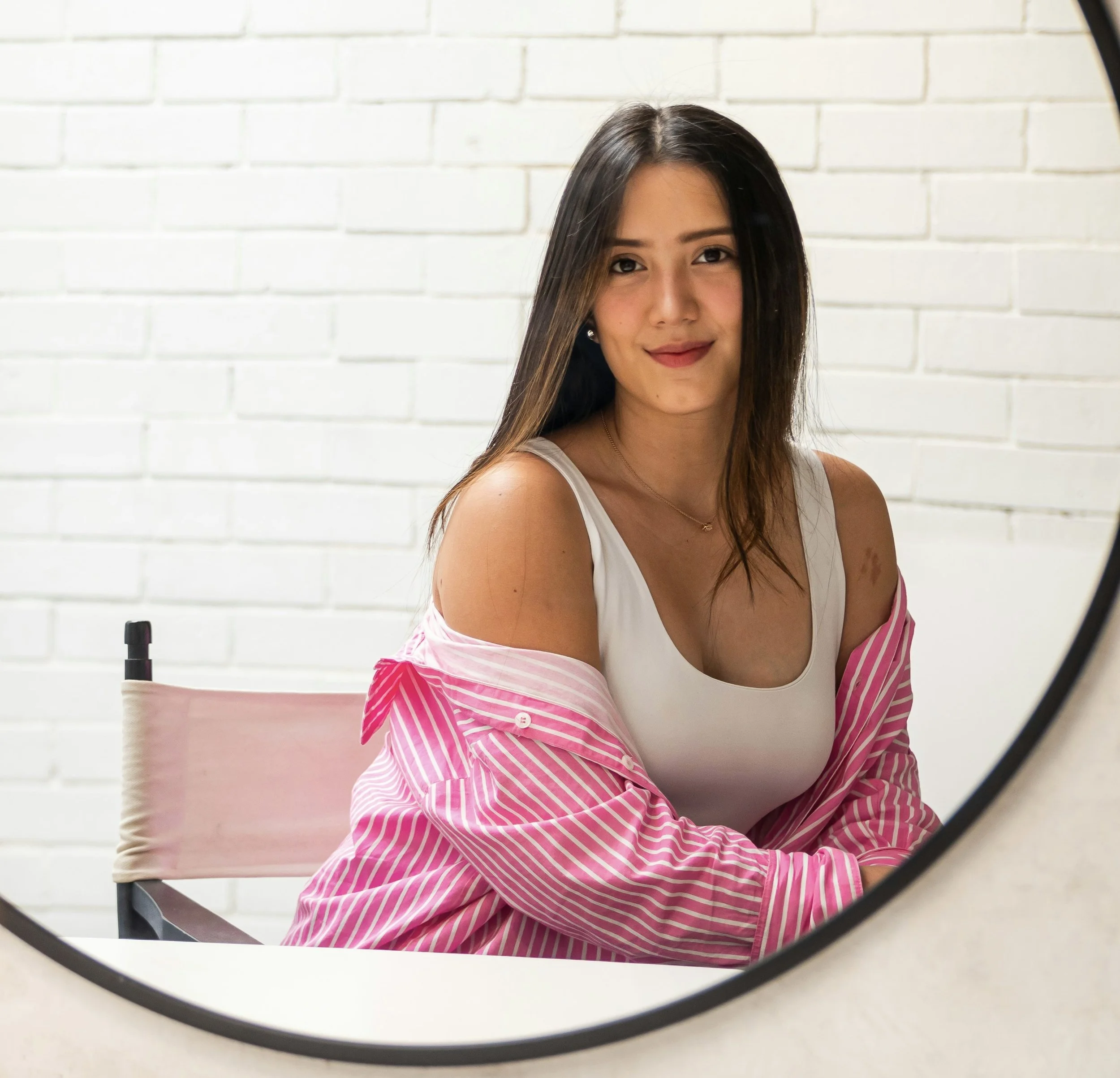 A young woman with long dark hair and light skin is taking a mirror selfie, wearing a white tank top and a pink and white striped shirt draped over her shoulders. She is sitting in front of a white brick wall.