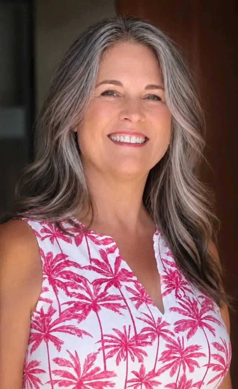 A smiling middle-aged woman with gray hair wearing a sleeveless white top with pink palm tree pattern, standing outdoors with a blurred background.