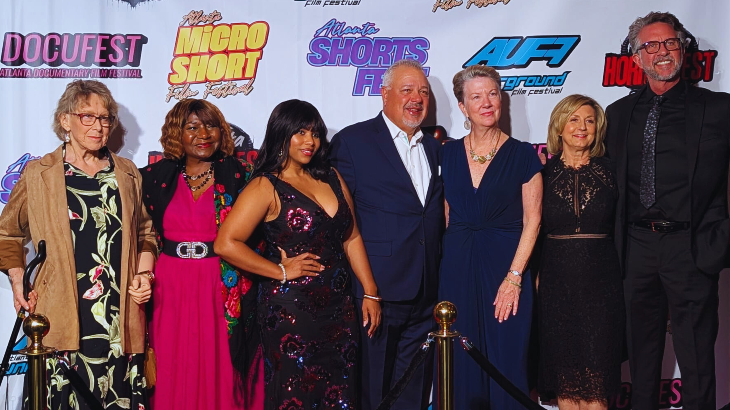 Group of seven people standing in front of a backdrop at the Atlanta Documentary Film Festival, posing for a photo. The backdrop displays festival logos including DOCUFEST, Micro Short, Shorts, AUF, and others. The individuals are dressed in formal and semi-formal attire.
