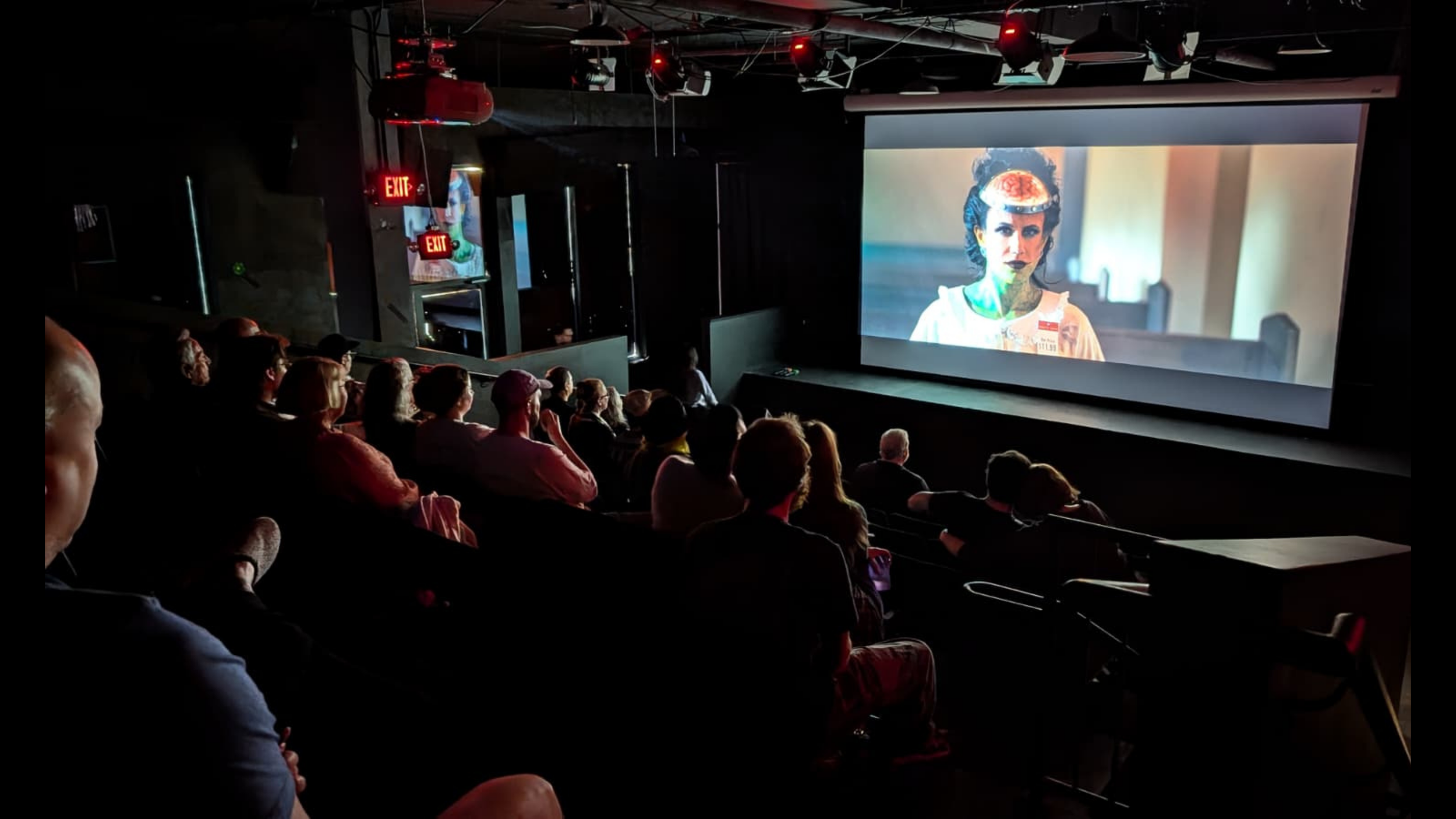 Audience watching a movie in a dark theater with a large screen showing a close-up of a woman with tattoos and traditional attire.