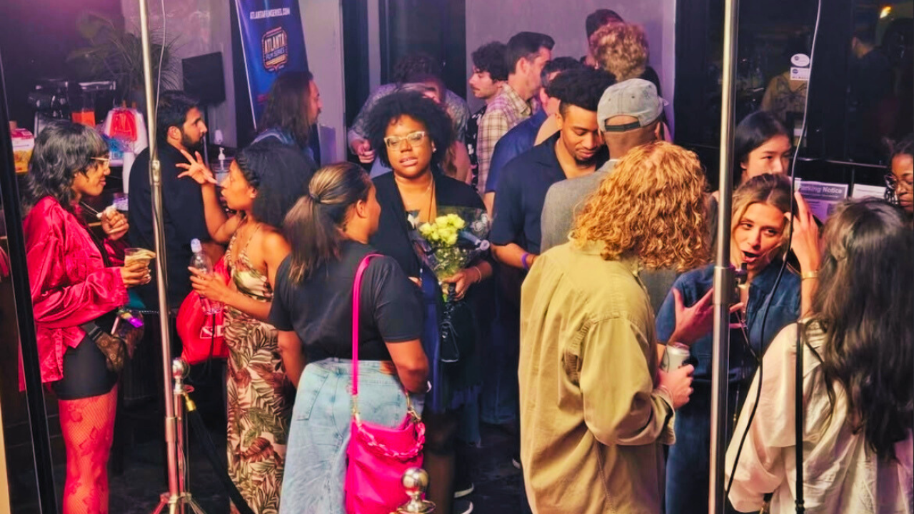 Crowd of diverse people waiting indoors, some talking, others with drinks or flowers, at an event or party.